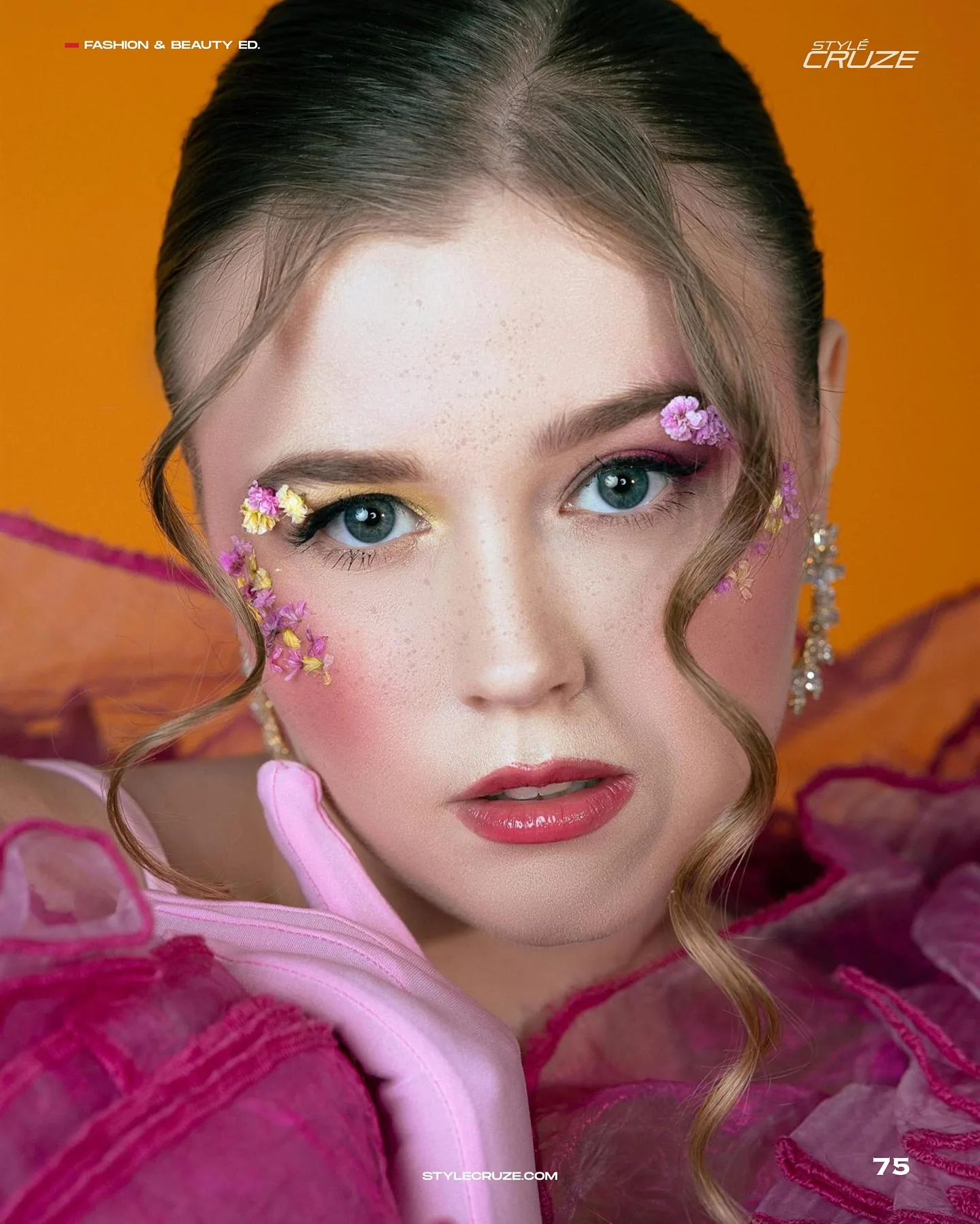 Beauty close-up on hot orange of a woman with slicked hair, floral appliqué eye makeup in pink and yellow, glossy coral lip and pink tulle at her shoulders.