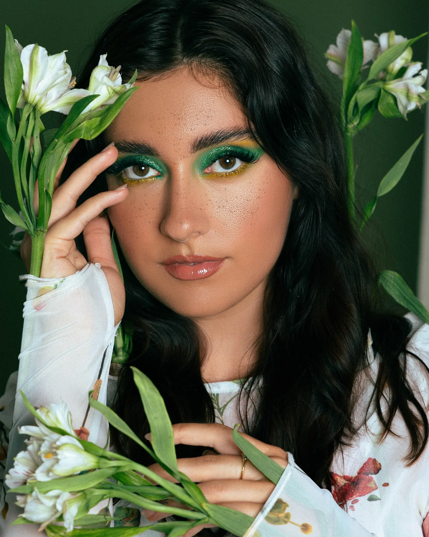 Beauty portrait of a dark-haired woman with emerald green eyeshadow in a sheer floral-print top, one hand raised to her brow, white alstroemeria blooms against a deep green wall.