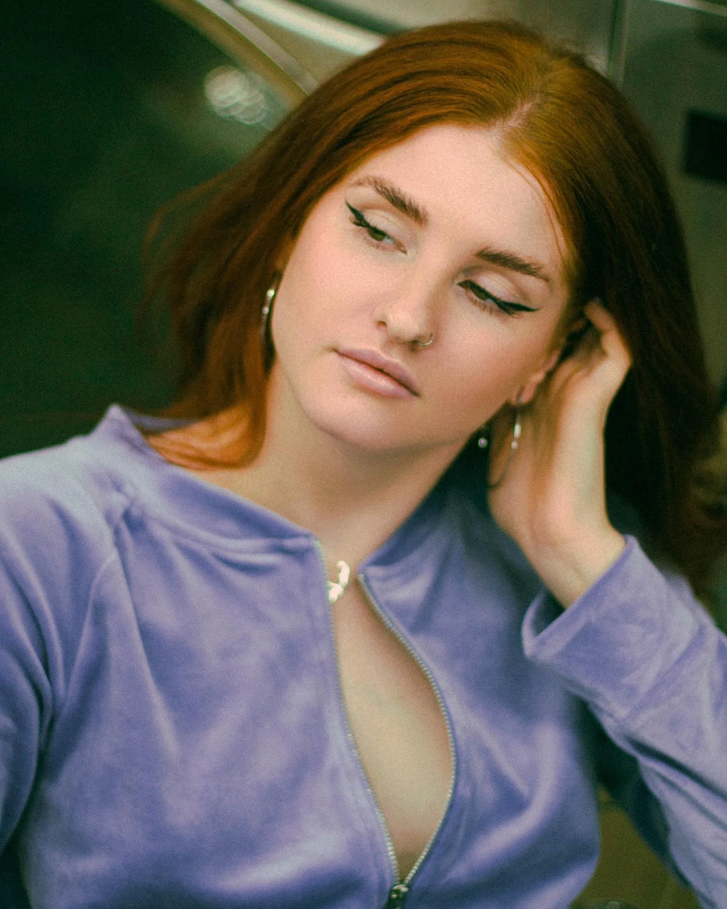 Close beauty portrait of a woman in a lilac velour zip-up, auburn hair falling forward, one hand pushed into her hair, eyes lowered, warm laundromat light behind her.