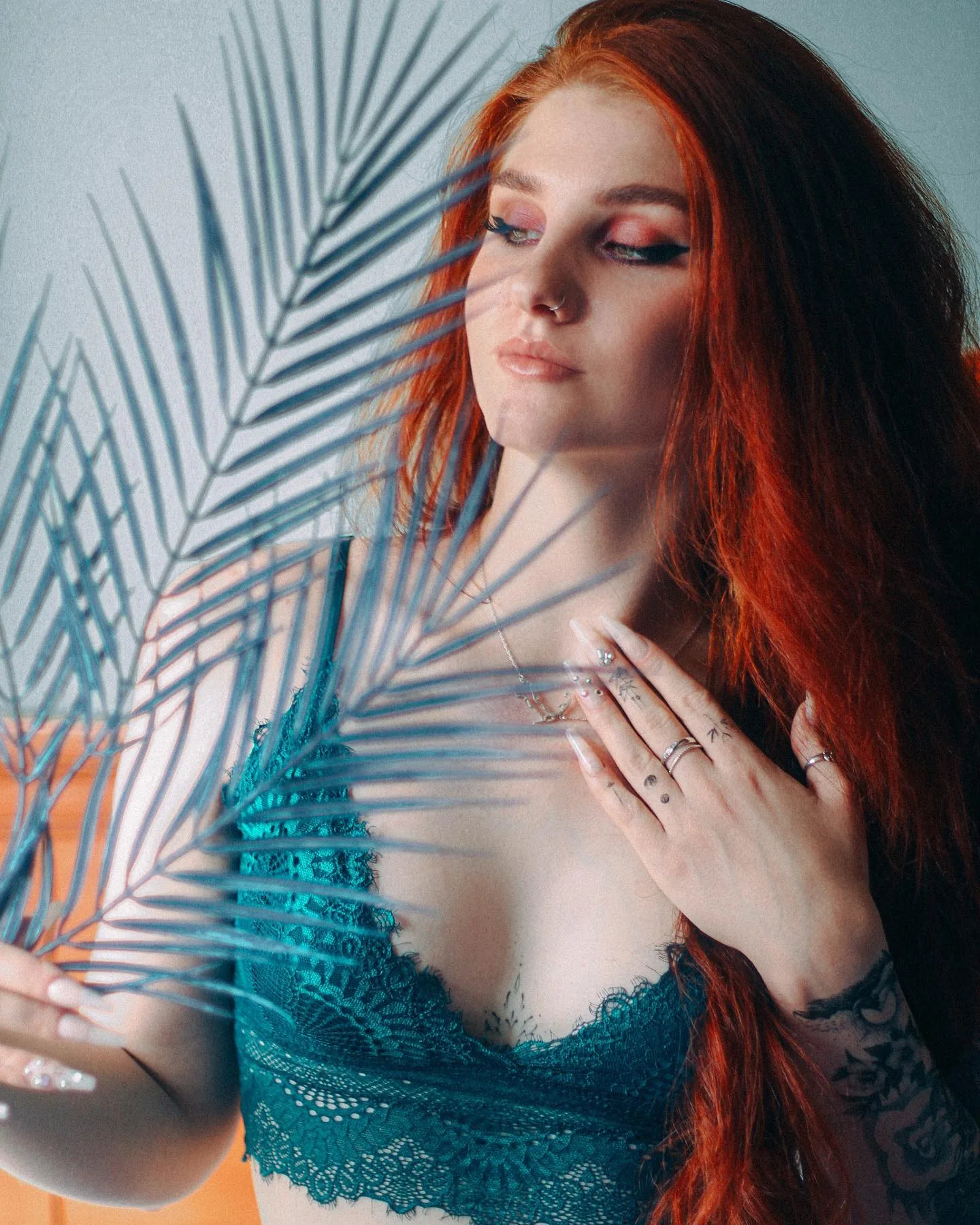 Woman with long copper-red hair in a teal lace bralette, eyes lowered, holding a blue-green palm frond across her chest against a pale grey wall.