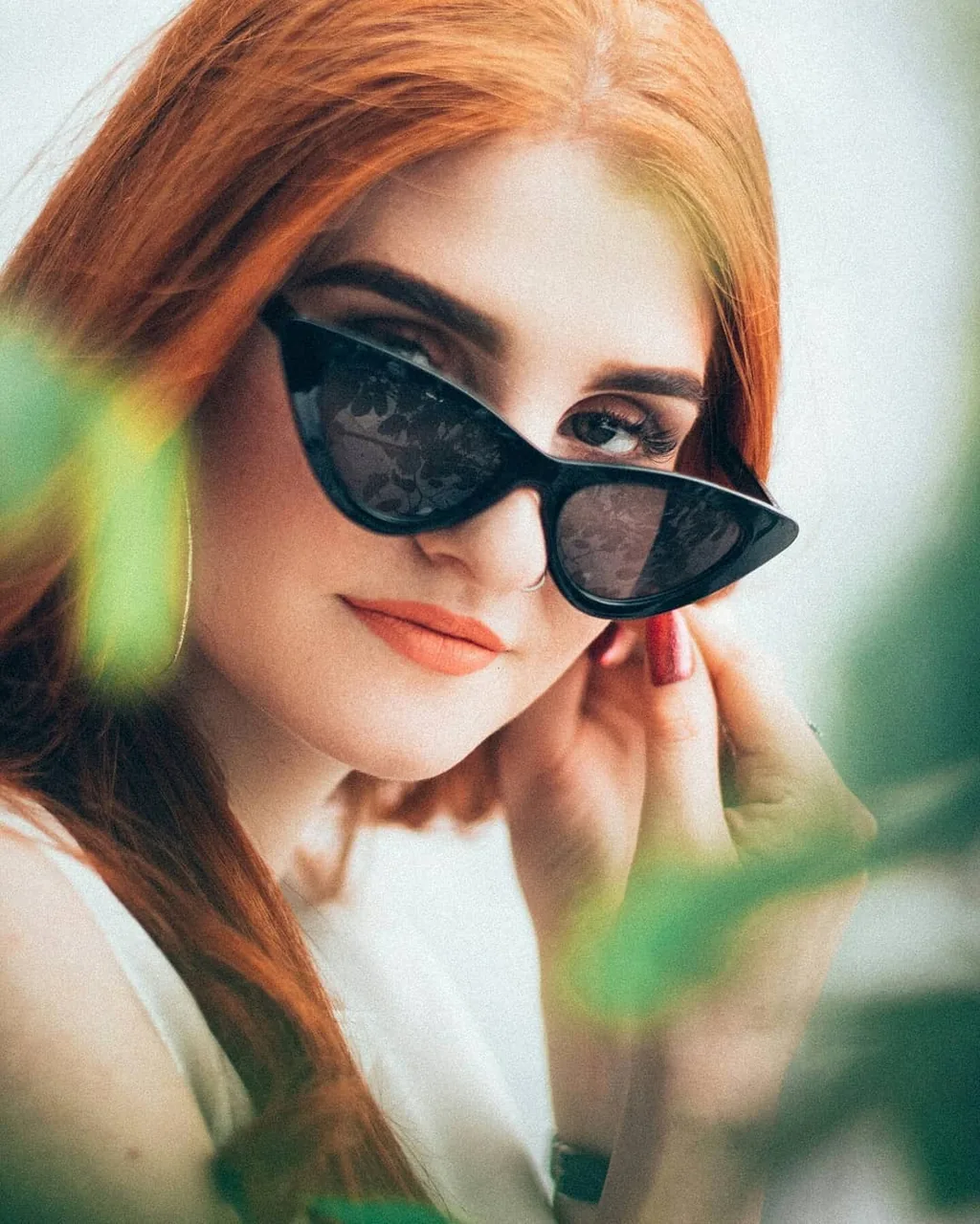Tight portrait of Daria in black cat-eye sunglasses pushed low, fingertips at her ear, gold hoop, coral lipstick, copper hair, shot through blurred green foliage in the foreground.