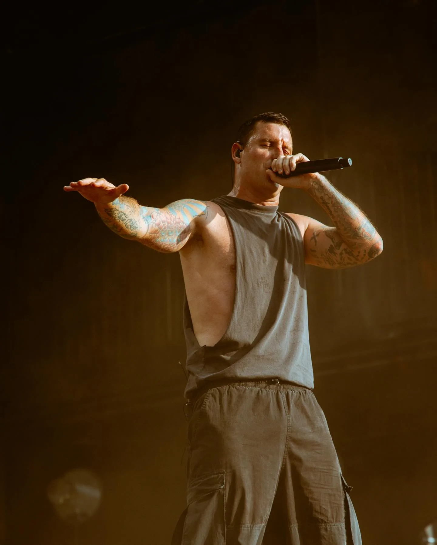 A tattooed singer in a grey sleeveless tank and cargo pants mid-performance, mic pressed to his mouth and one arm flung out to the side, isolated against warm golden stage haze and darkness