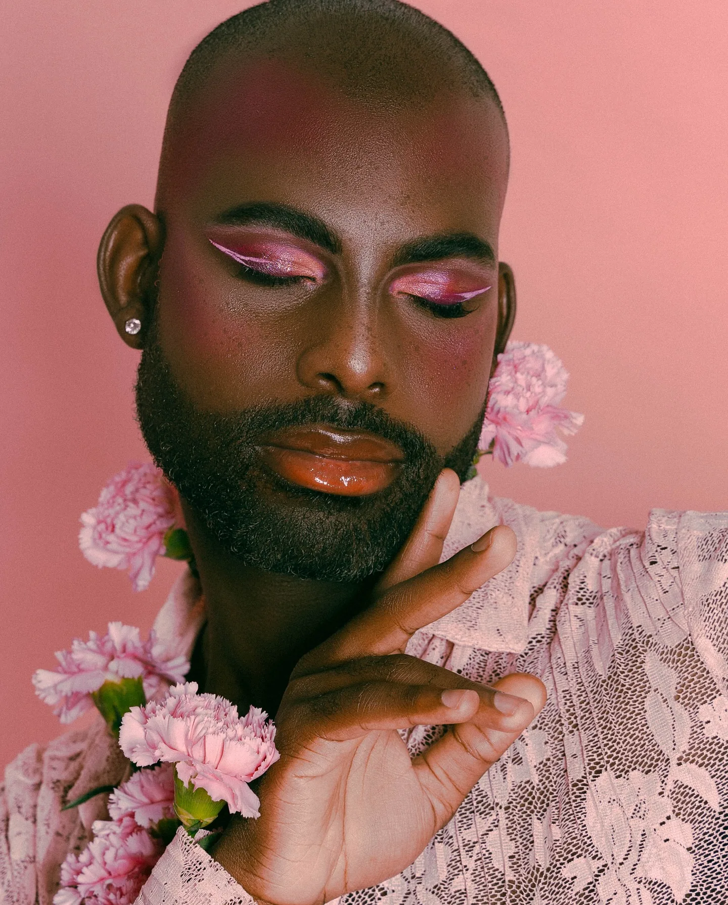 Close-up of a bald bearded Black man in a sheer pink lace shirt against a pink backdrop, wearing bright pink and orange eyeshadow and glossy lips, eyes closed, pink carnations tucked at his shoulder