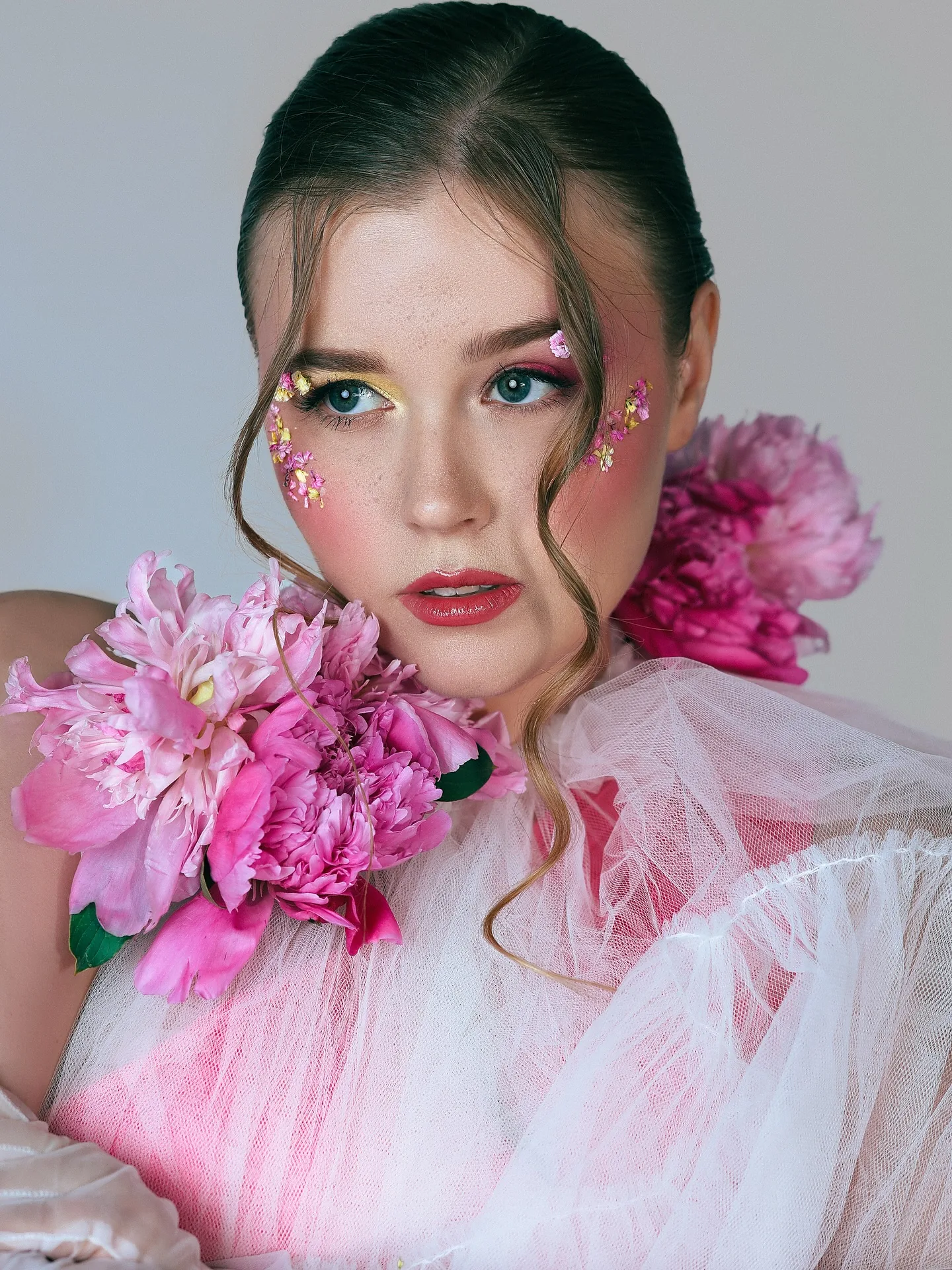 Beauty close-up of a model in pale pink tulle with an armful of pink peonies at her shoulder, floral glitter appliqué scattered around her eyes, hair slicked back