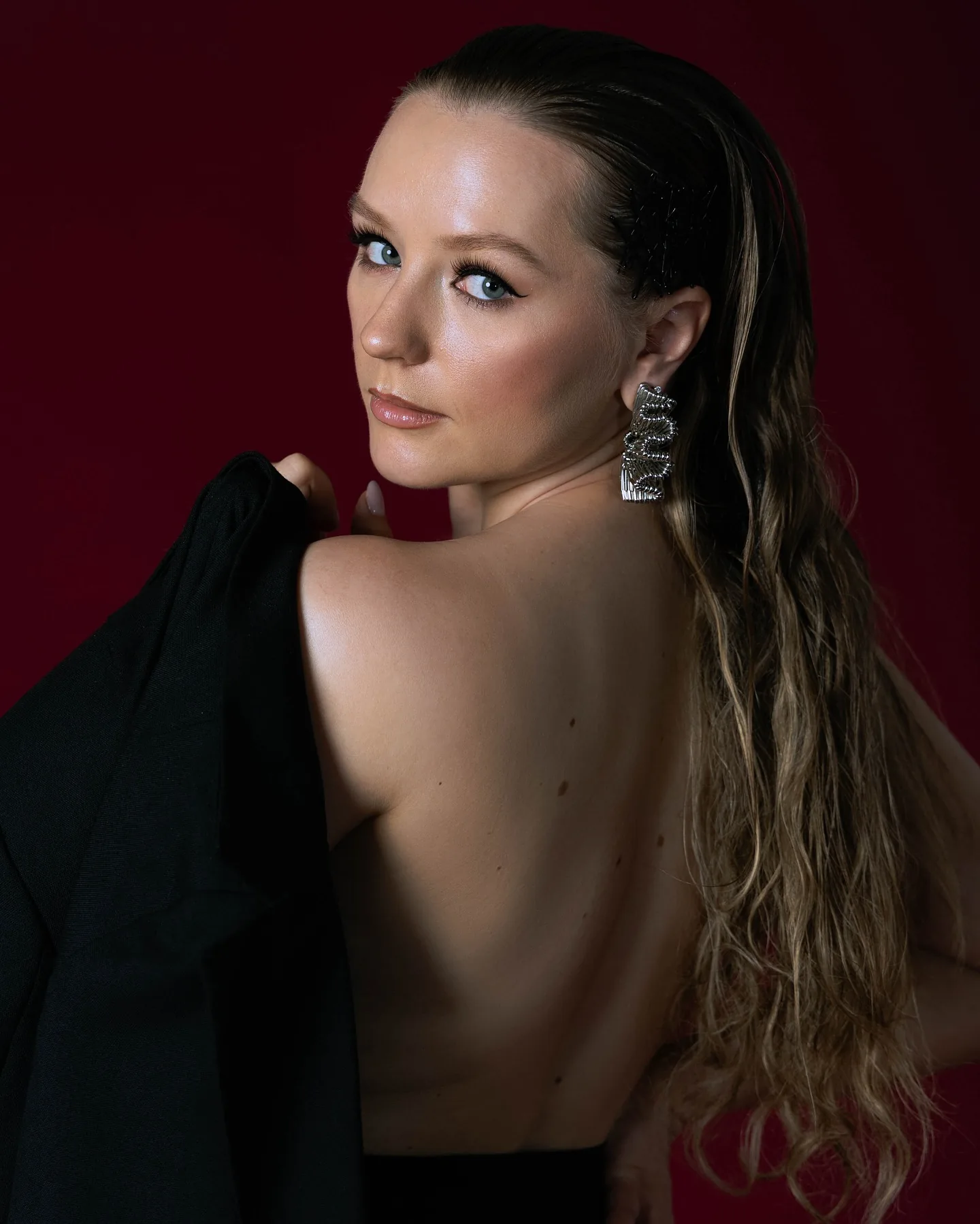 A blonde model on a deep crimson backdrop with a black blazer slipped off her bare shoulders and back, turning to look over her shoulder into the lens with long crystal earrings catching the light
