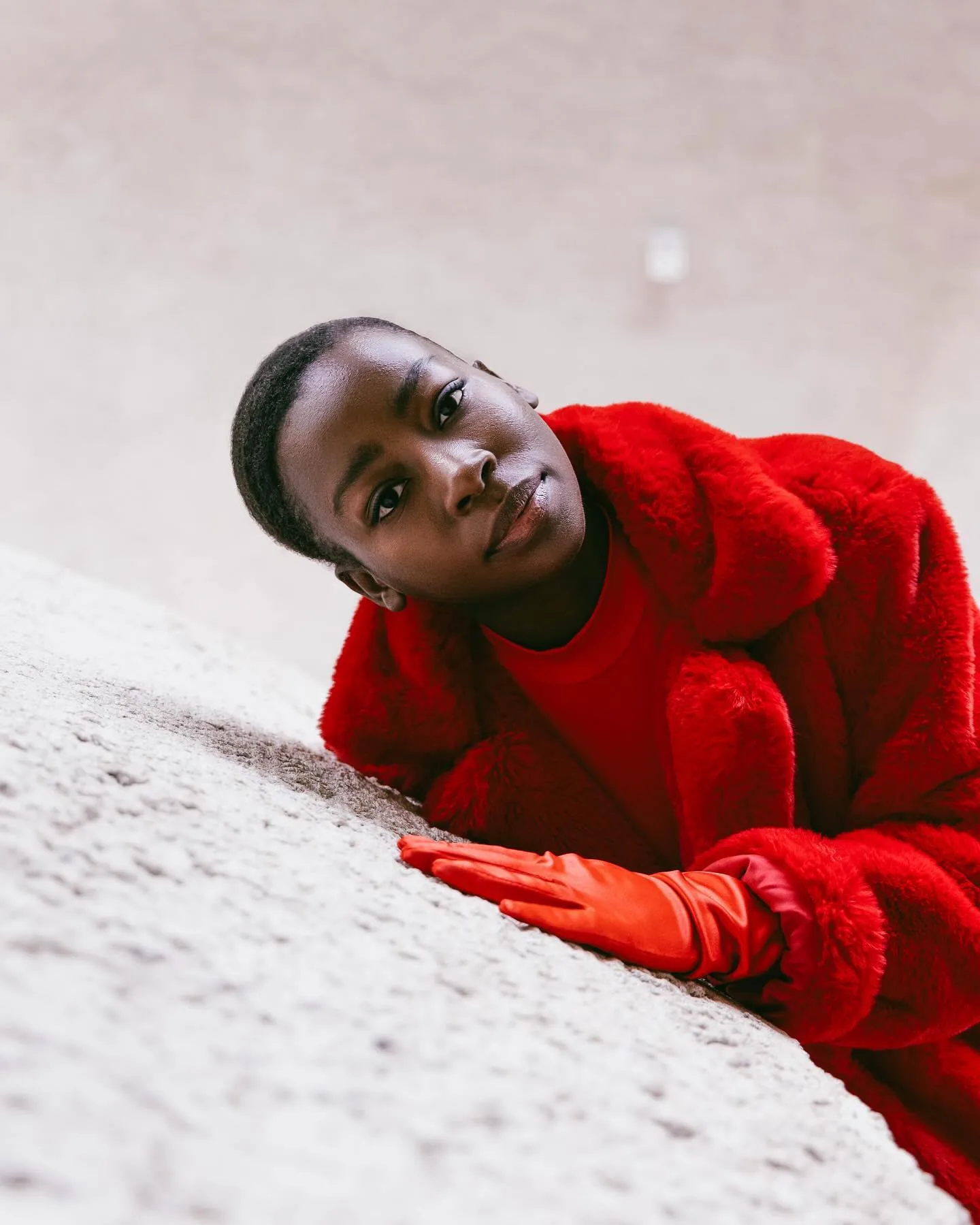 A model in a red fur coat, red turtleneck and red satin gloves leans back against a curved white concrete wall, head tilted, looking directly into the lens