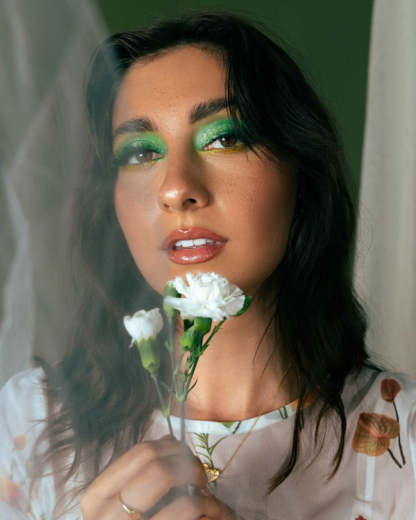 Close-up of a dark-haired model in emerald green graphic eyeshadow behind a veil of sheer white tulle, holding a single white carnation near her chin against a deep green wall