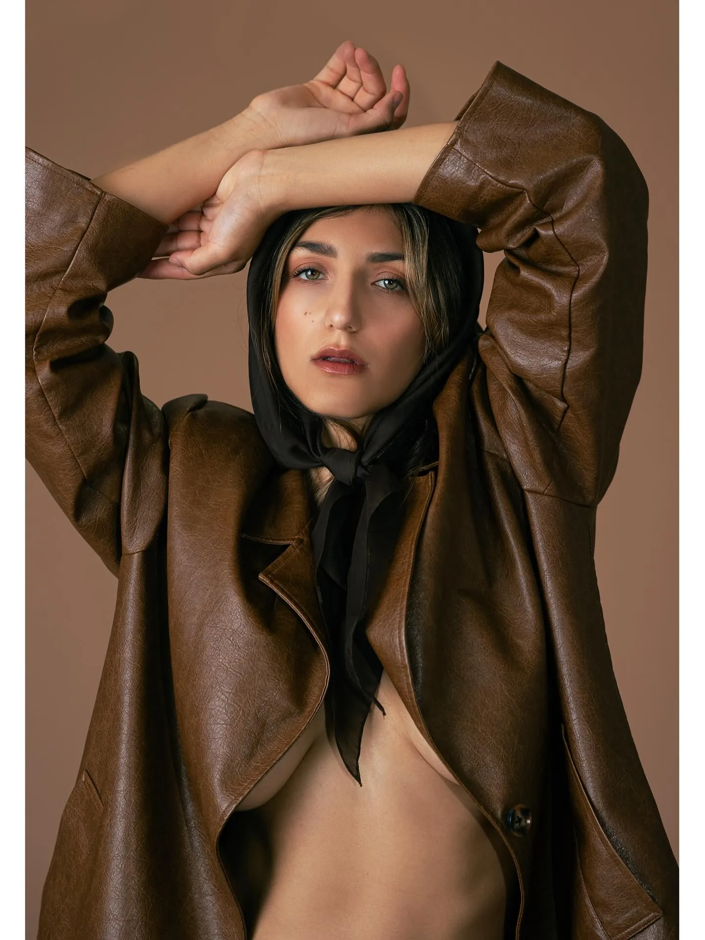 A dark-haired model in an oversized brown leather jacket and black headscarf against a warm tan seamless, one arm raised and folded over her head, gaze level with the camera