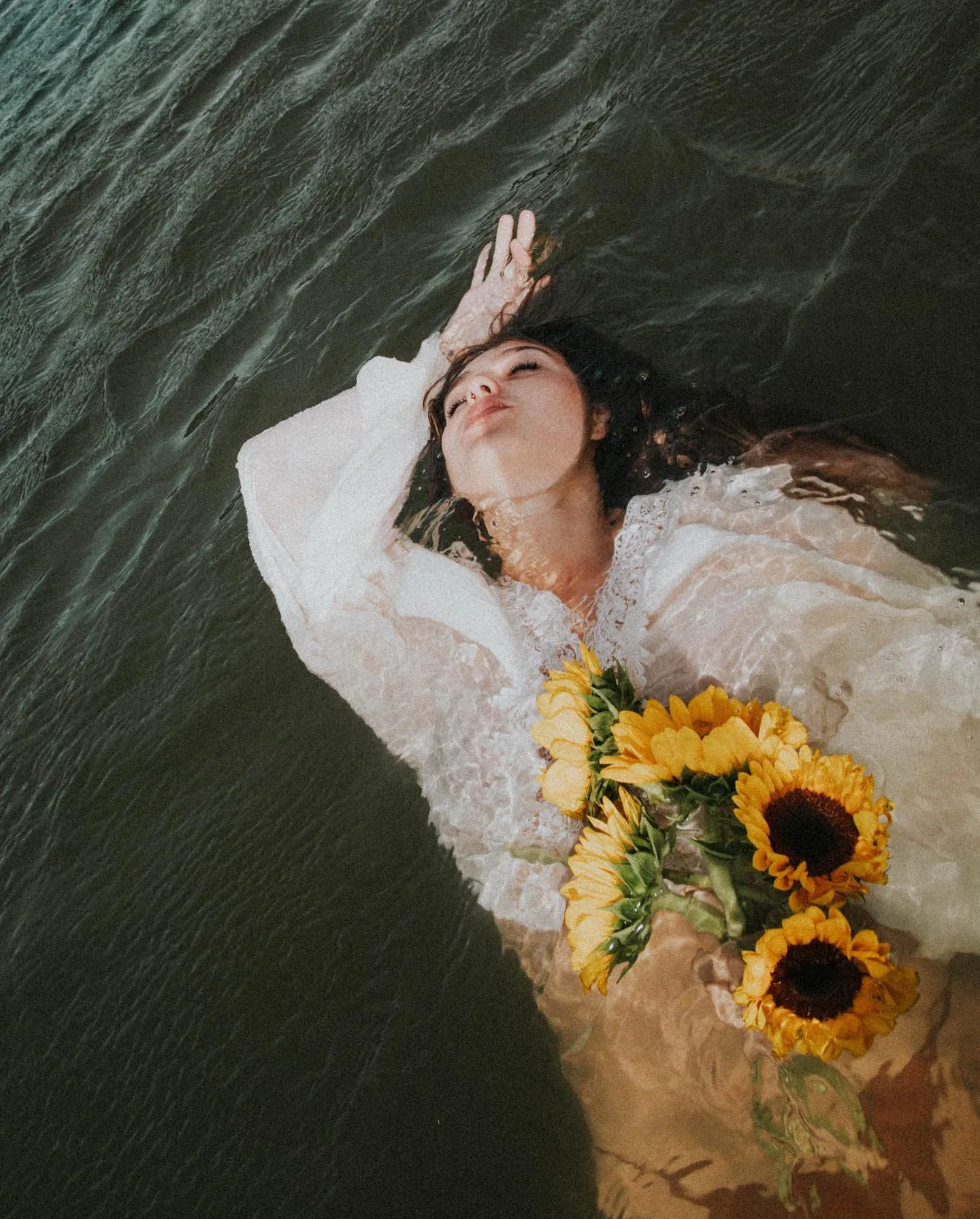 A woman in a white lace dress floats on her back in dark green water, eyes closed and one arm curled above her face, with a bunch of yellow sunflowers resting across her chest just under the surface