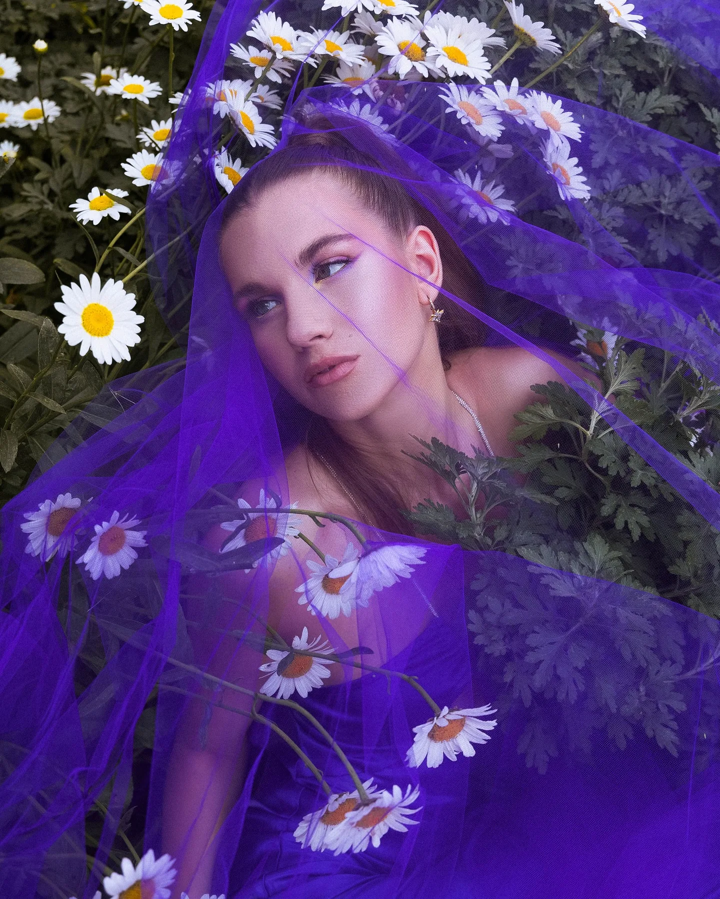 A model lies in a bed of white daisies wrapped in electric-violet tulle that veils her face and shoulders, looking sideways past the camera with lilac eyeshadow and a small star earring