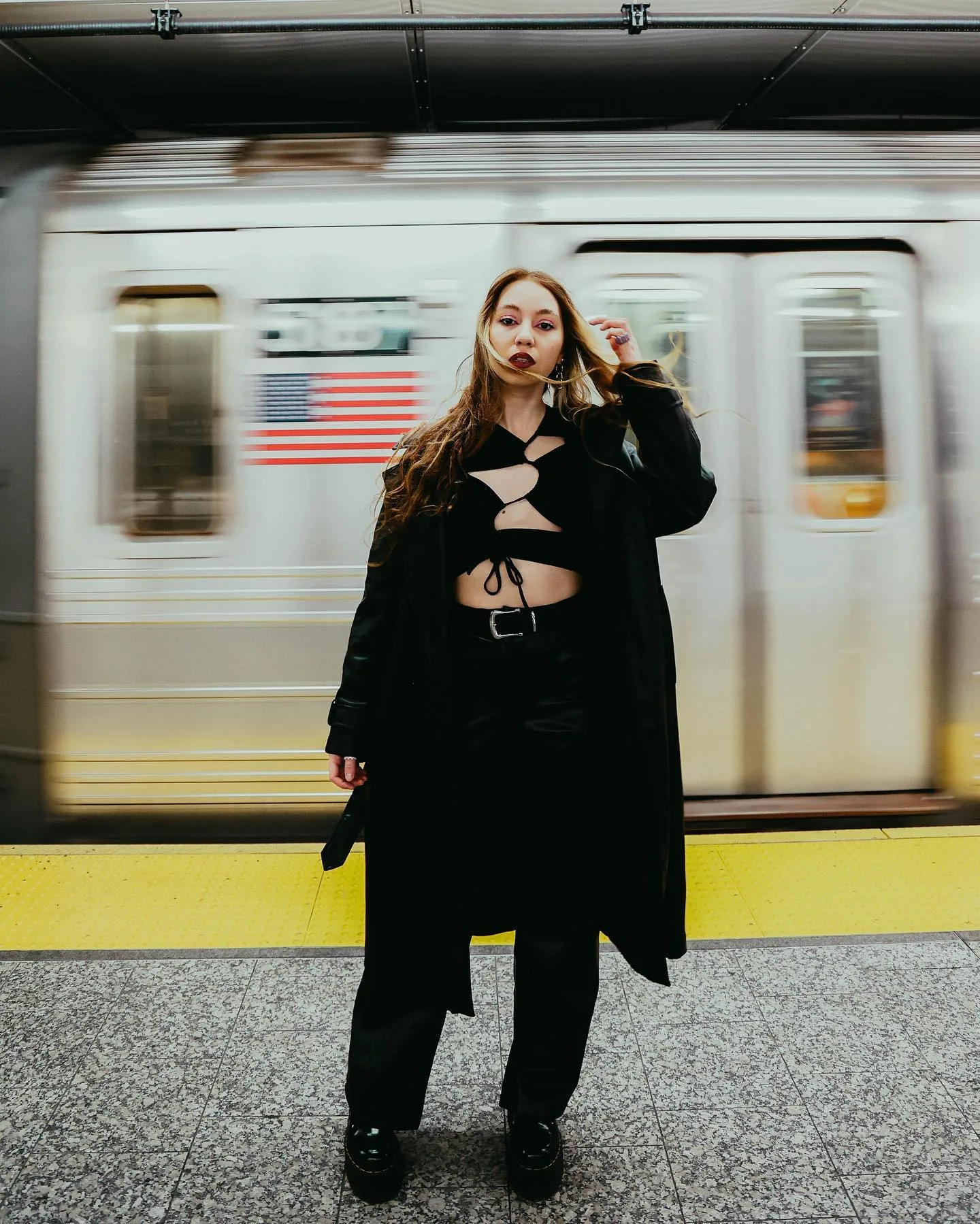 A woman in a long black coat stands still on a subway platform while a train streaks past behind her in motion blur, an American flag decal smeared across its side