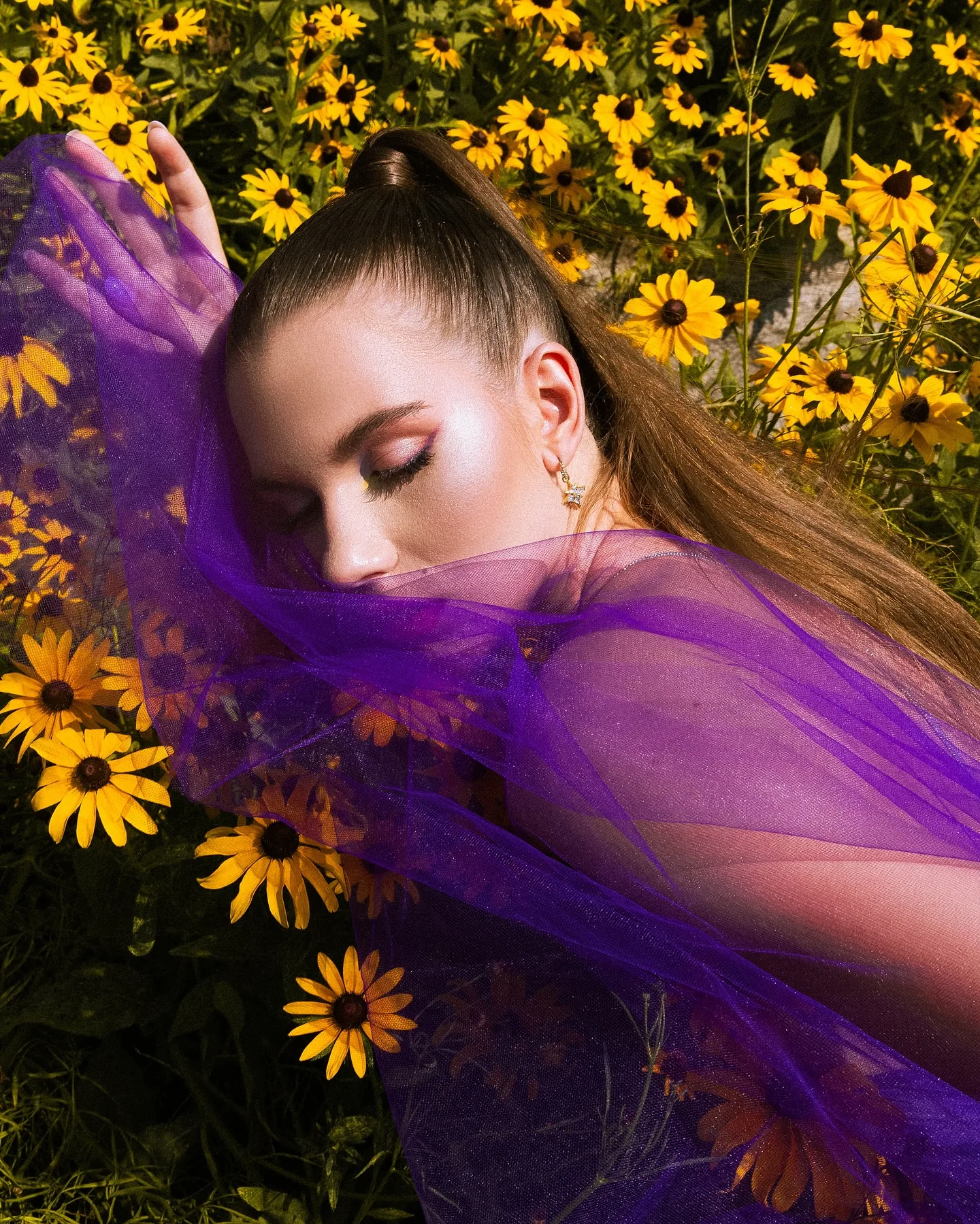 A woman lying back in a bed of yellow black-eyed Susans, wrapped in translucent violet tulle that sweeps diagonally across the frame, eyes closed, hair in a high ponytail, hard sunlight on her skin