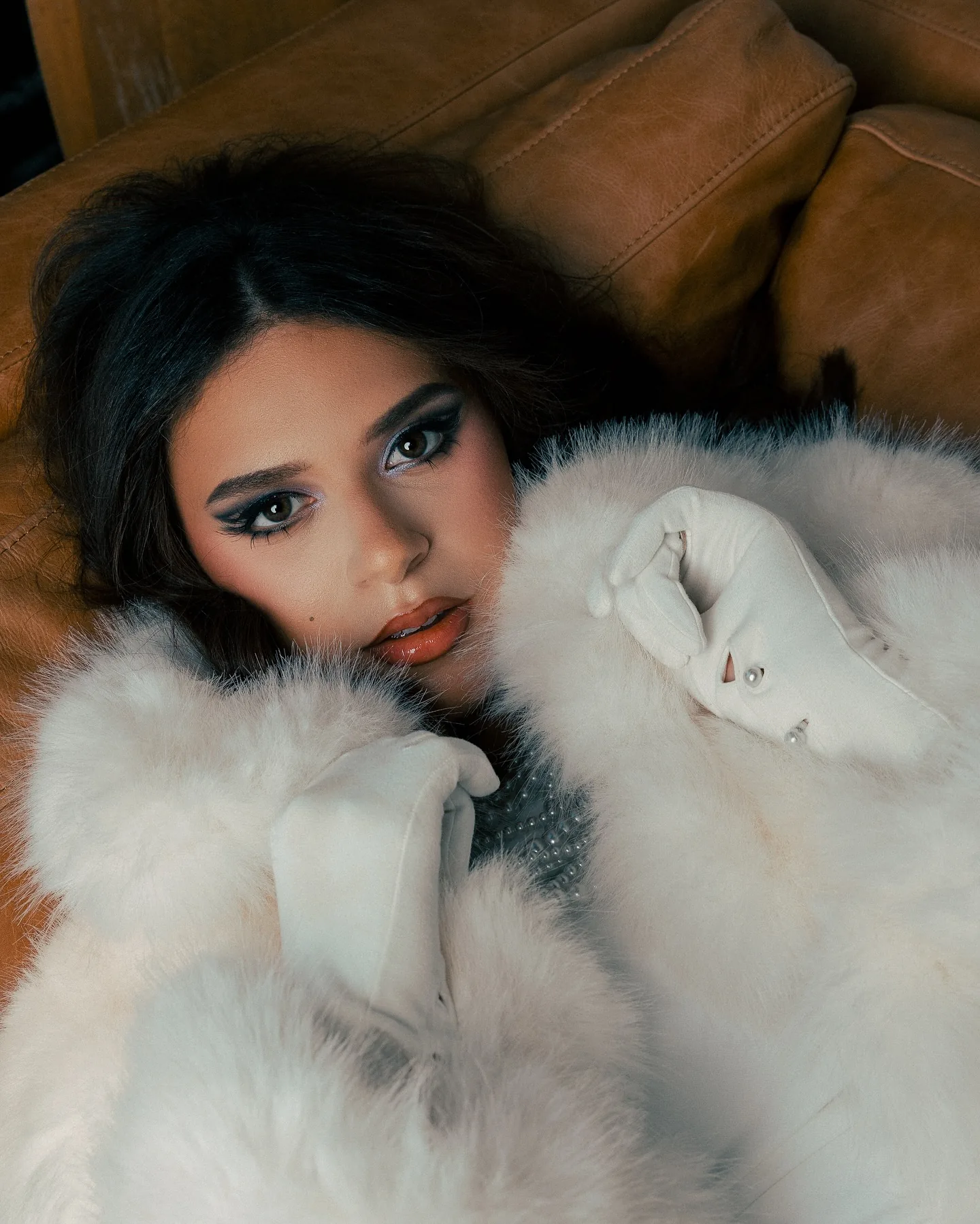 Overhead close-up of a dark-haired woman lying on a tan leather sofa in a white fur coat, wearing dramatic eye makeup and deep red lipstick, a strand of pearls at her collar