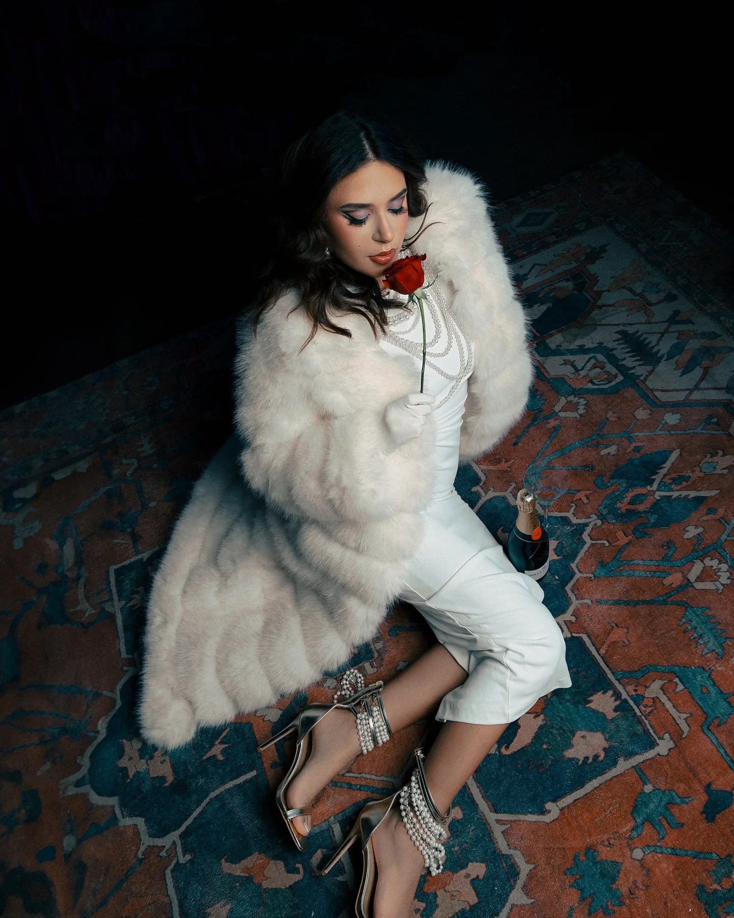 Overhead full-body shot of a woman in a white fur coat and white trousers reclining on a red and blue Persian rug, gold strappy heels, a red rose held at her mouth