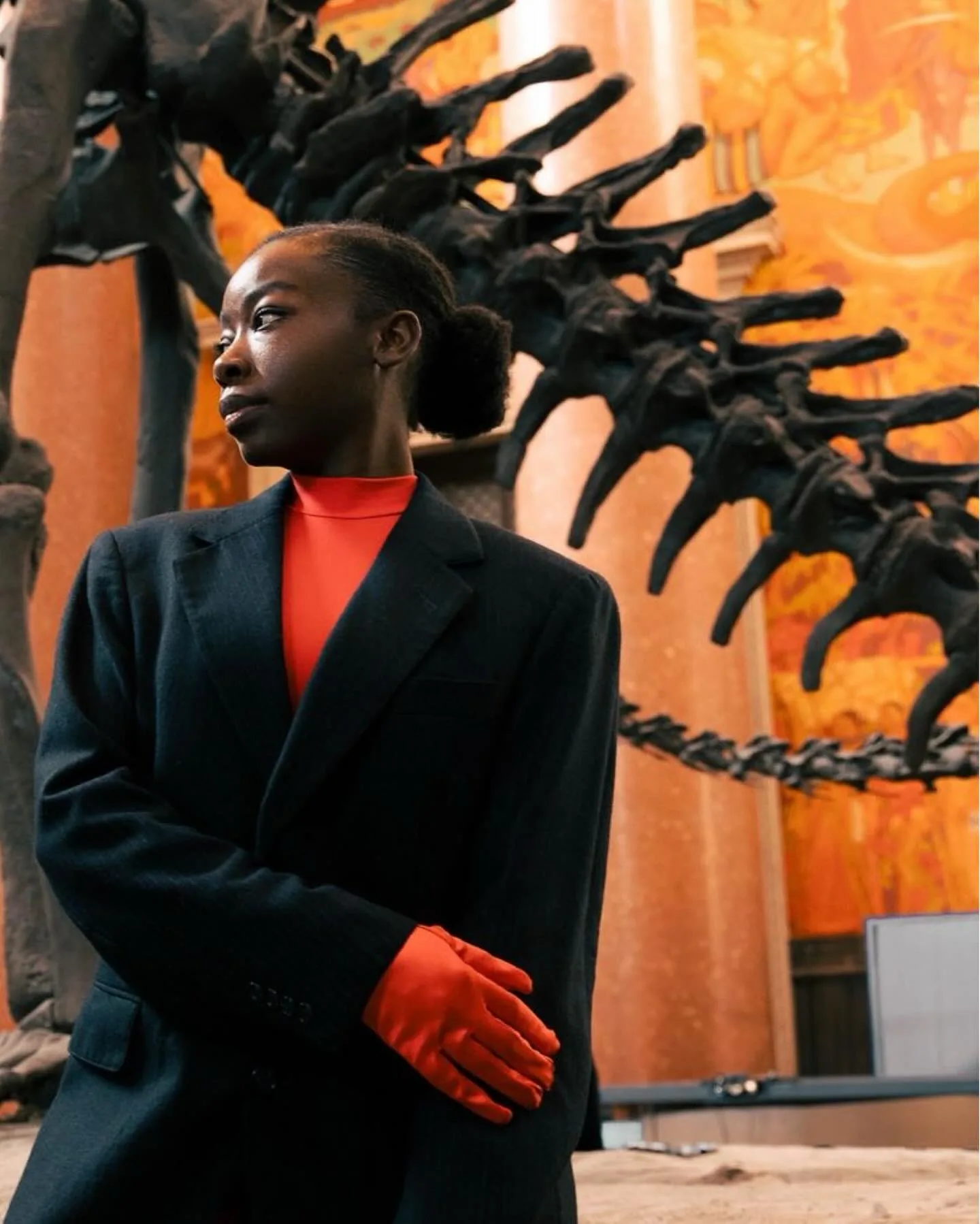 A model in an oversized black blazer, red turtleneck and red satin gloves stands in profile beneath the black vertebrae of a mounted dinosaur skeleton, an orange museum mural glowing behind her