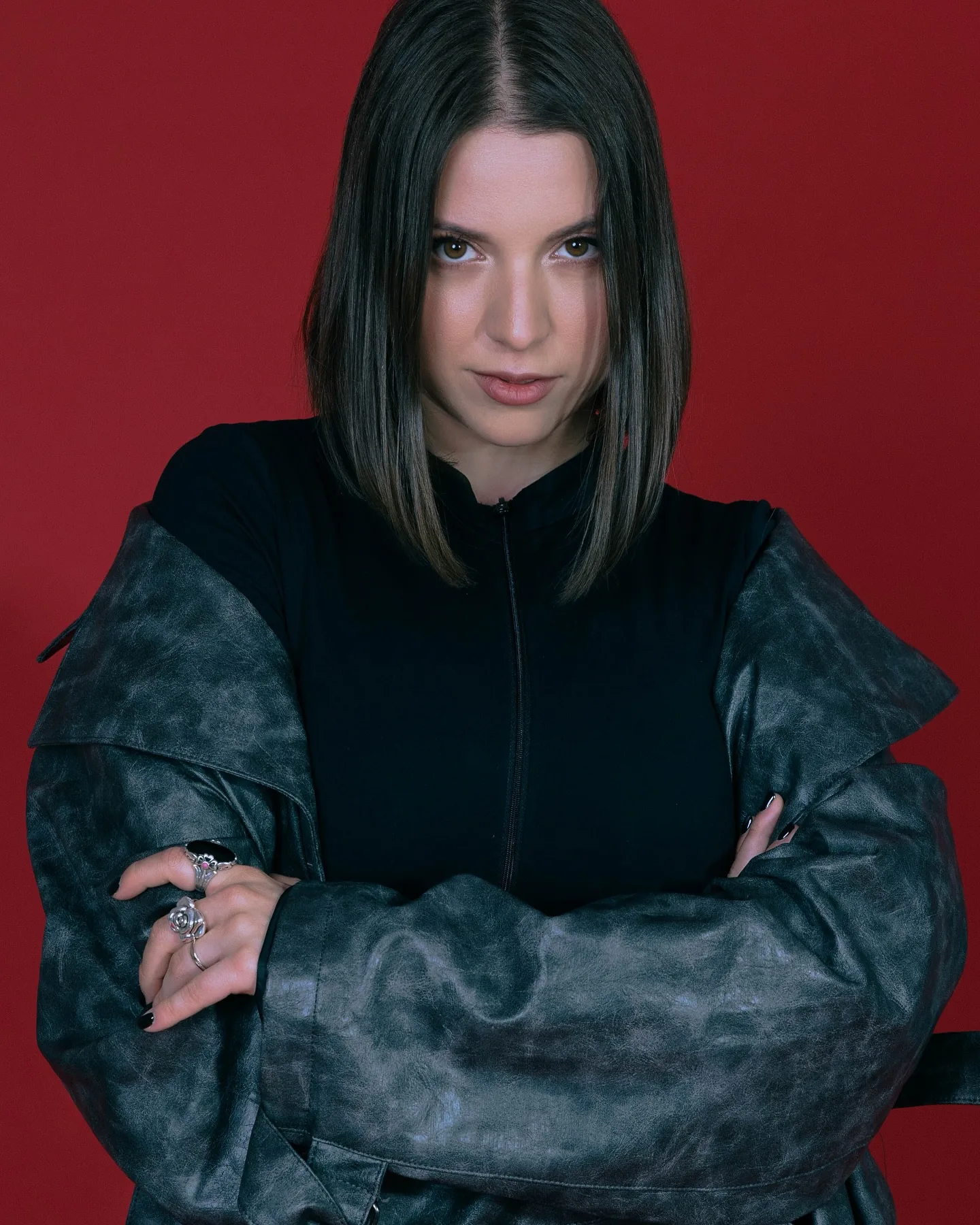 A model with a sharp dark bob stands arms crossed on a red backdrop in a structured teal-grey coat with padded sleeves worn over a black top, staring level into the camera