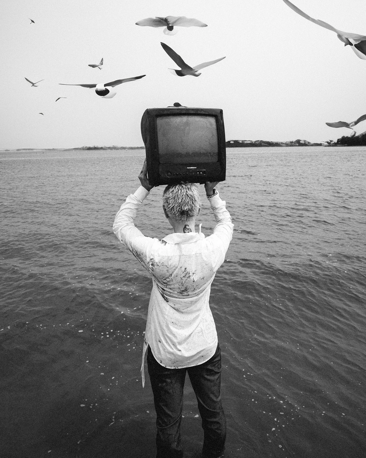 Black-and-white frame of a person in a white shirt standing waist-deep in the sea, lifting a vintage CRT television above their head, gulls scattered across the pale sky behind them