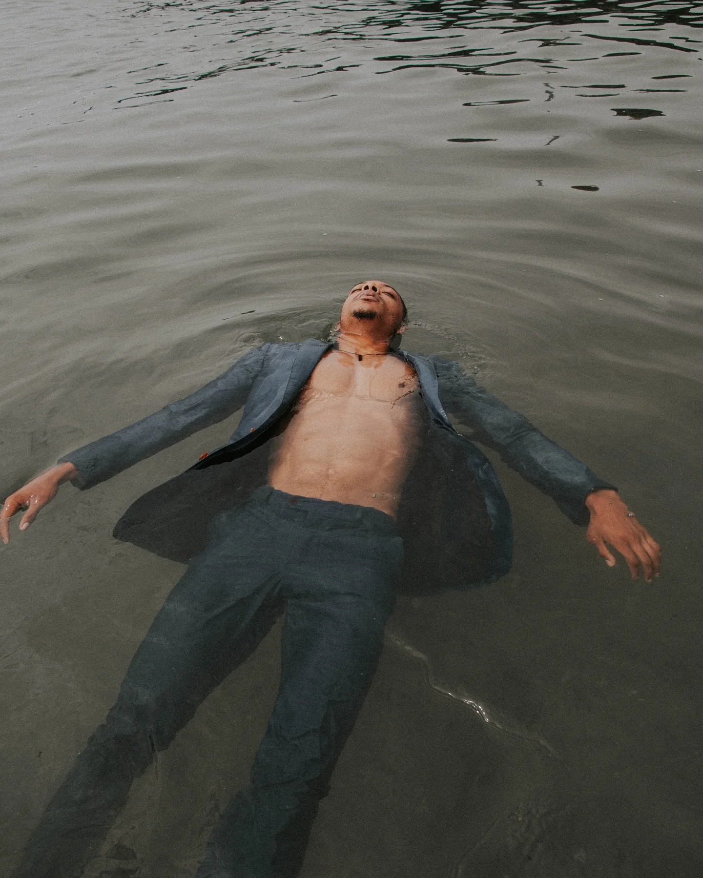 A man in a soaked slate-blue suit, jacket open over a bare chest, floats on his back in still grey-green water, arms spread and face tilted up with eyes closed, ripples radiating out around his head