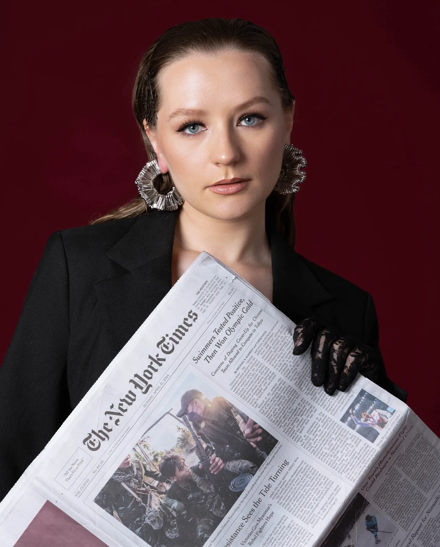 Studio editorial on crimson: a model in a black blazer and black lace gloves holds an open copy of The New York Times across her body, chin level, silver fan earrings framing her face