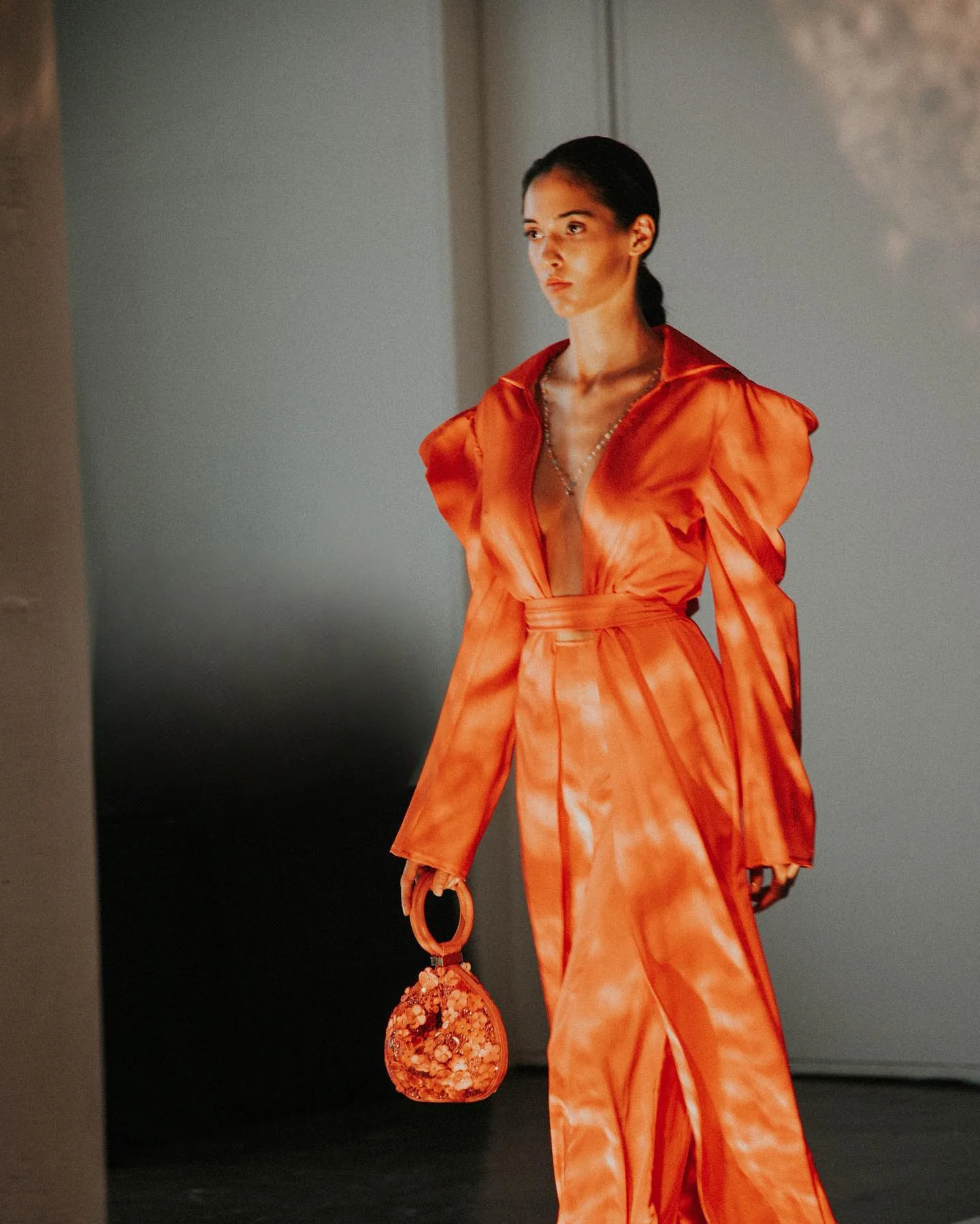 A model in a vivid orange satin gown with exaggerated puffed shoulders and a deep plunging neckline stands in dappled light against a pale grey wall, holding a small beaded orange top-handle bag