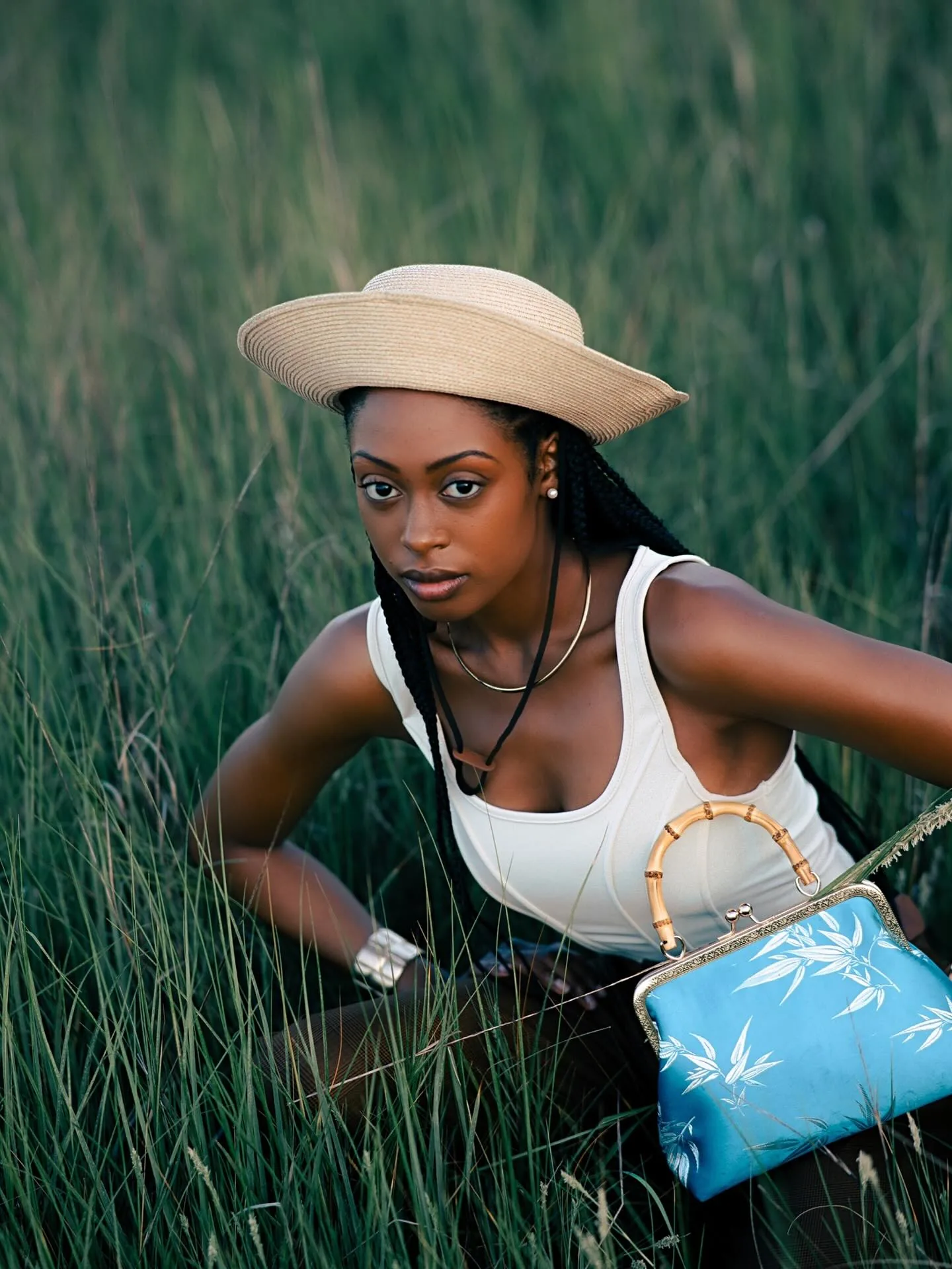 A Black woman in a wide cream straw hat and white tank top crouching in tall green marsh grass at dusk, one hand planted in the grass, holding a sky-blue bamboo-handled handbag, looking straight into the lens