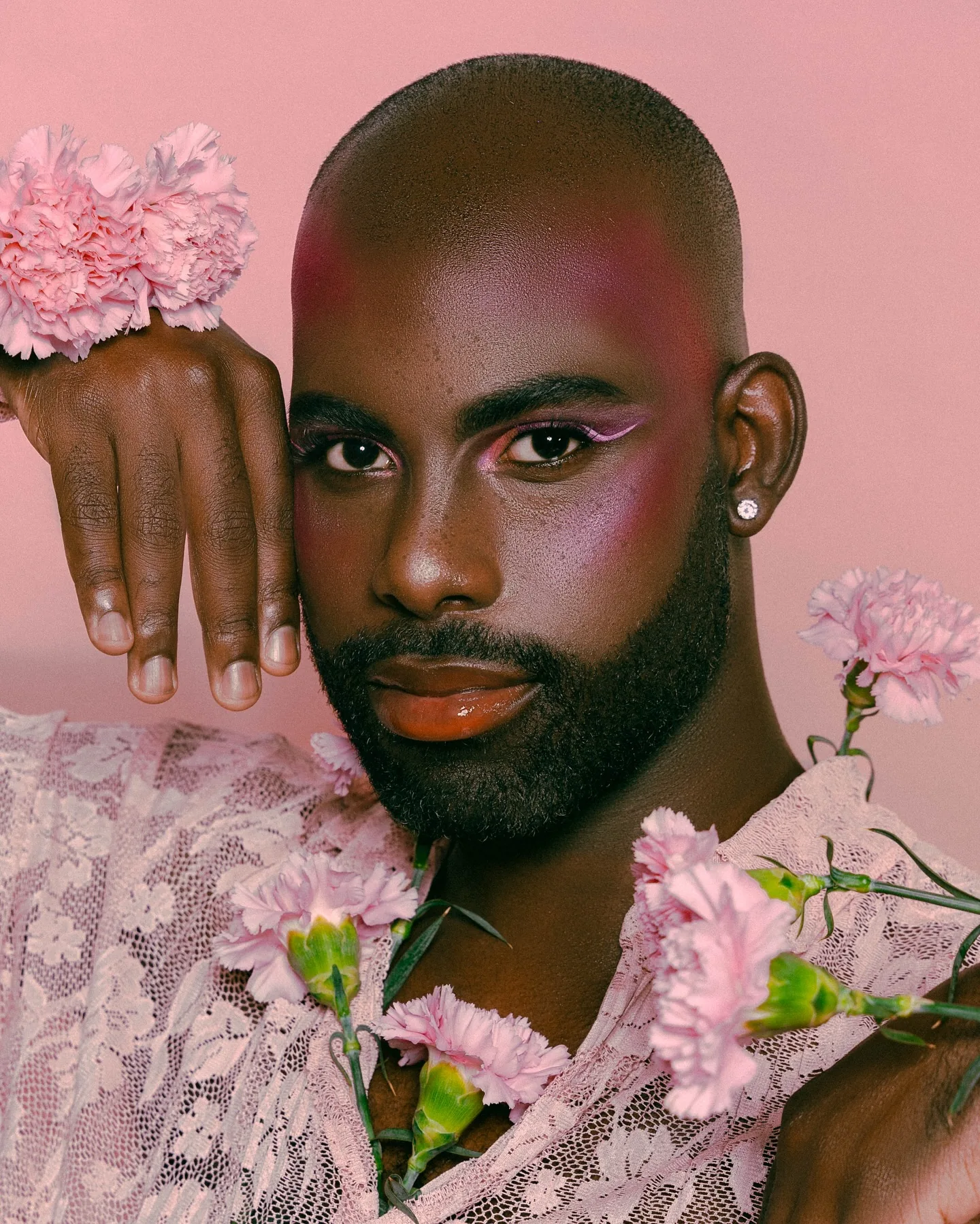 A bearded man in a sheer pink lace shirt on a soft pink set, pink eyeliner and gloss, one hand raised beside his head with pale carnations tucked at his wrist and collar