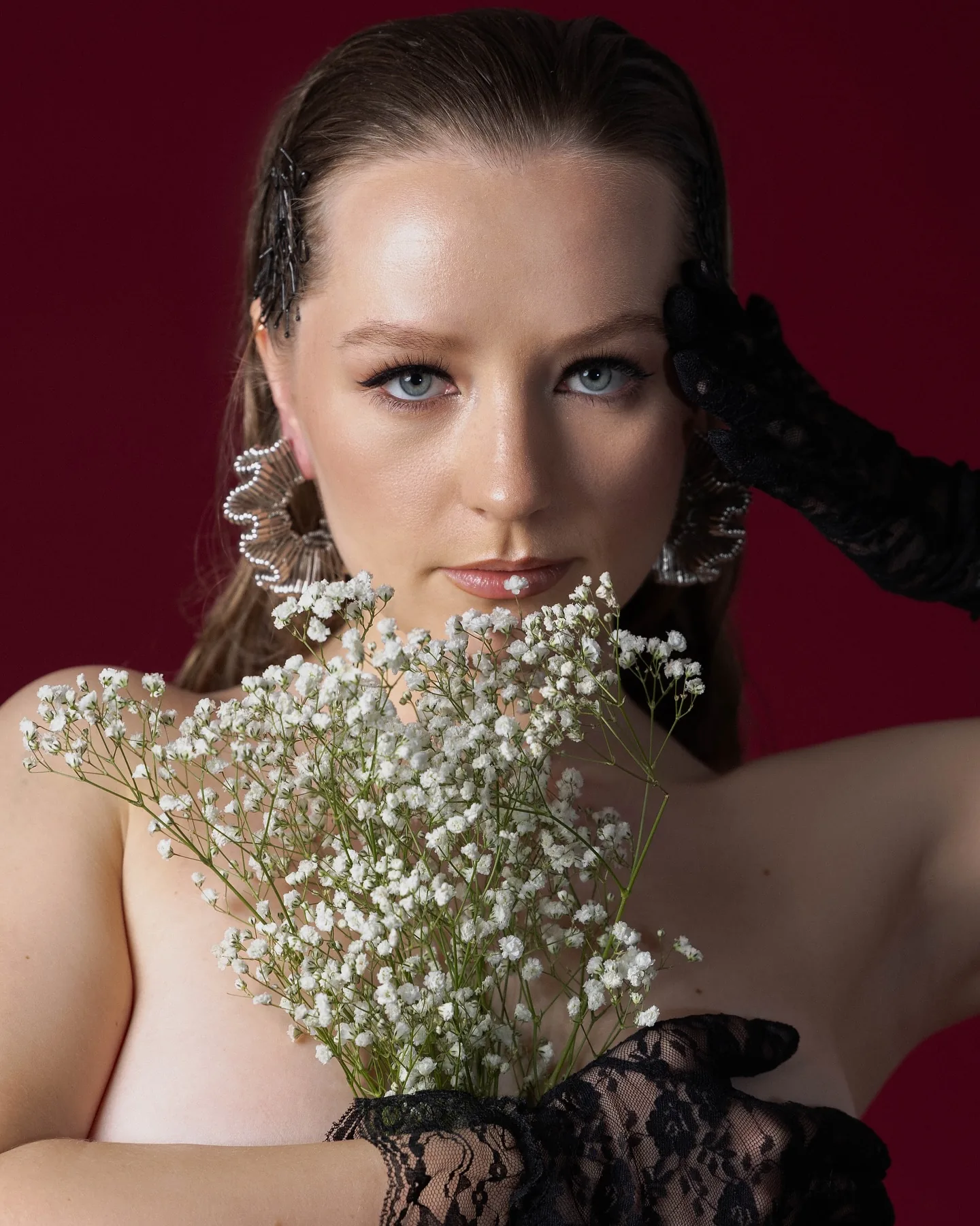 Beauty portrait on crimson: a model with slicked-back hair and black lace opera gloves holds a spray of white baby's-breath across her bare chest, one gloved hand at her temple, gazing straight ahead