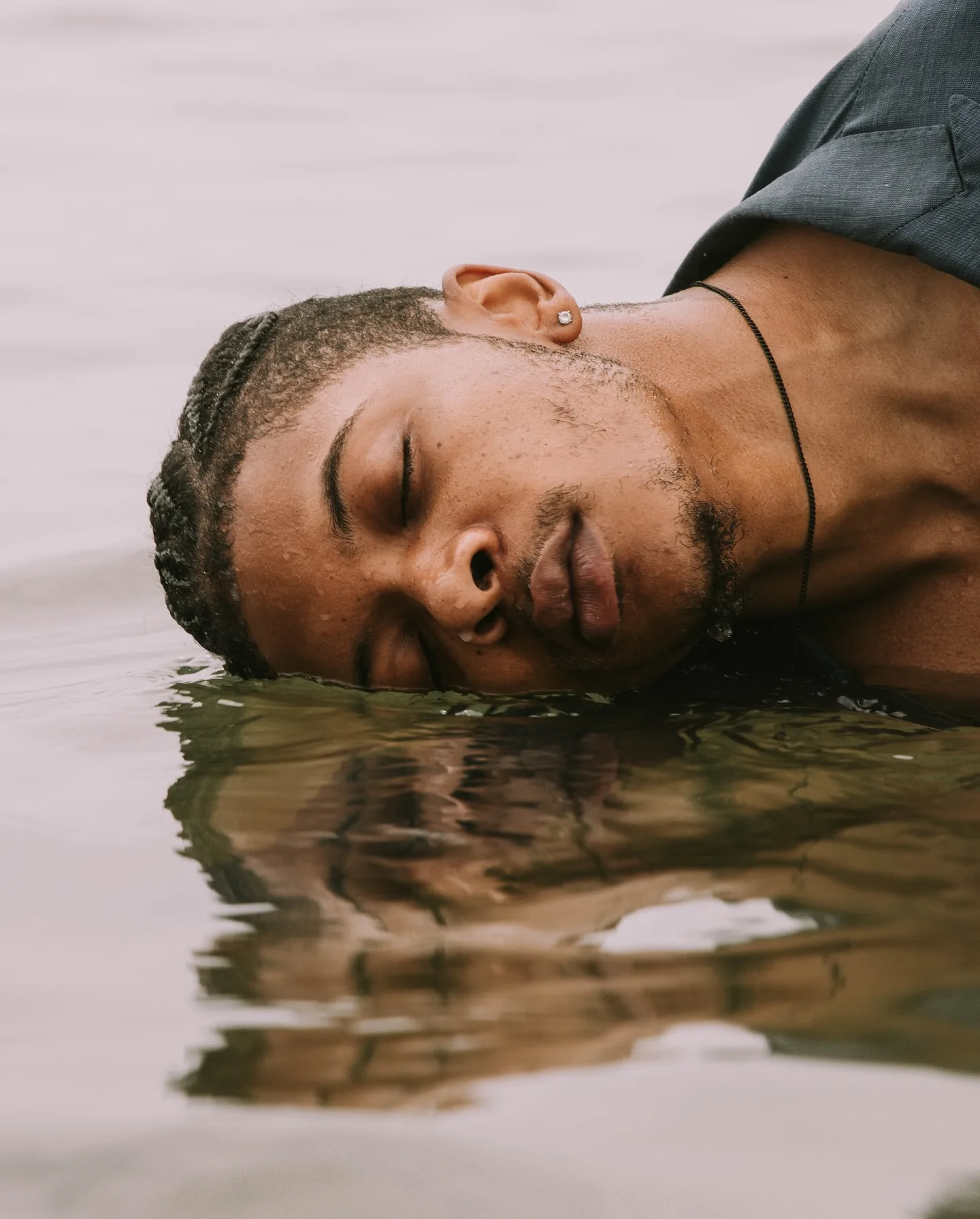 Extreme close-up of a man's face resting sideways at the surface of pale water, eyes closed and mouth parted, his reflection rippling below him and a wet blue suit shoulder at the top of the frame