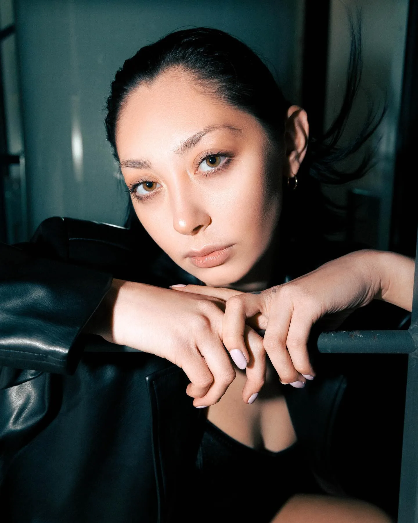 Night portrait of a dark-haired woman in an oversized black leather coat, forearms folded over a metal scaffolding rail with her chin above her hands, staring straight down the lens