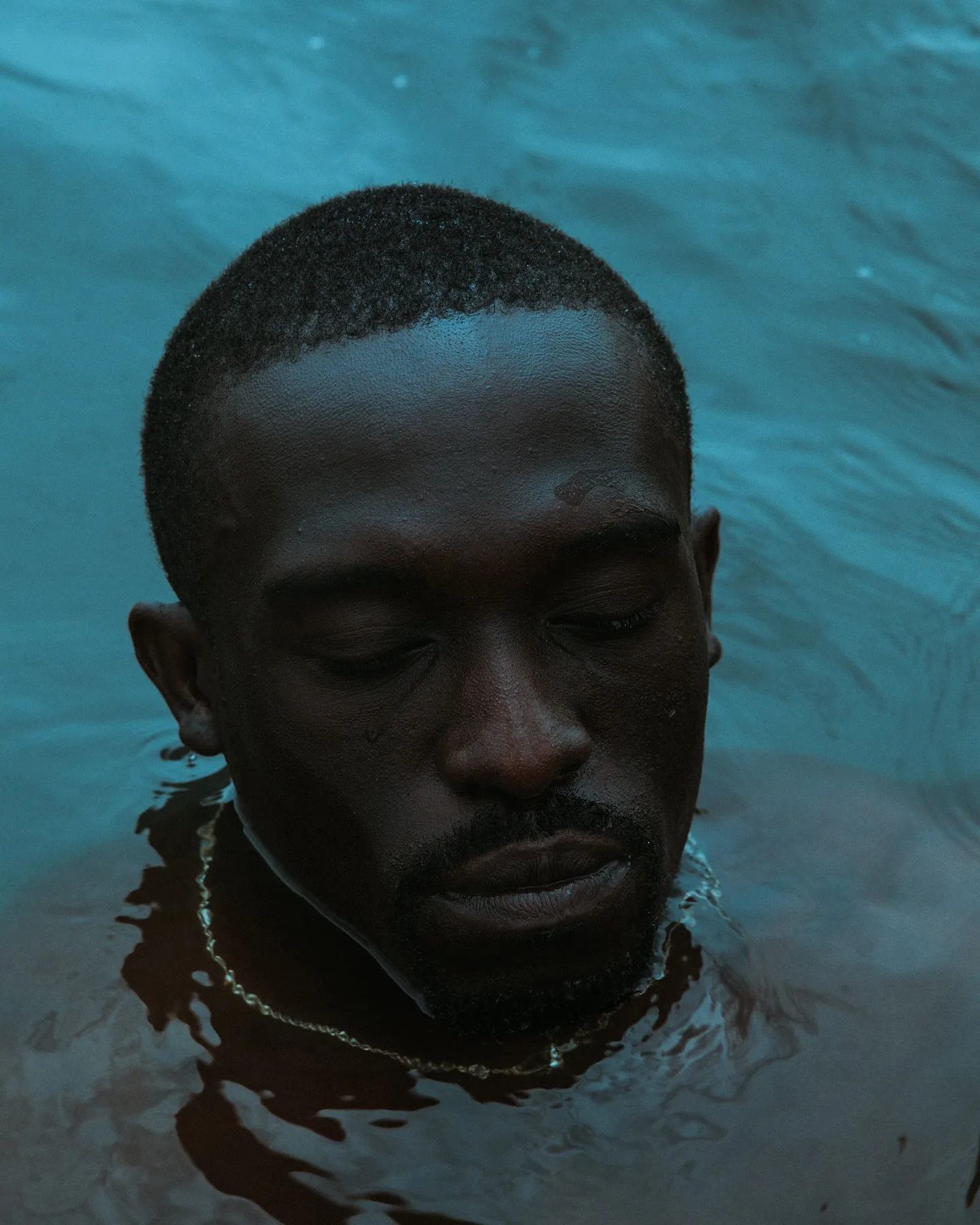 A Black man submerged to his chin in still teal-green water, eyes closed and head tilted slightly down, a thin gold chain visible just beneath the rippling surface, water filling the entire frame