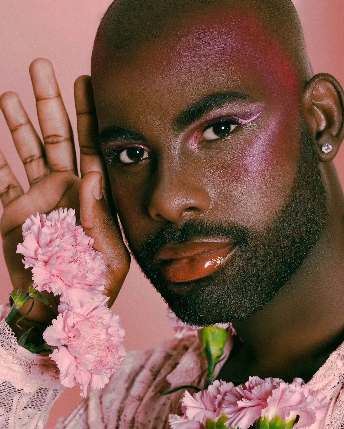 Tight beauty portrait of a bearded man against pink, wearing pink graphic eyeliner and gloss, one open palm pressed beside his face while pale pink carnations crowd in at his jaw and lace sleeve