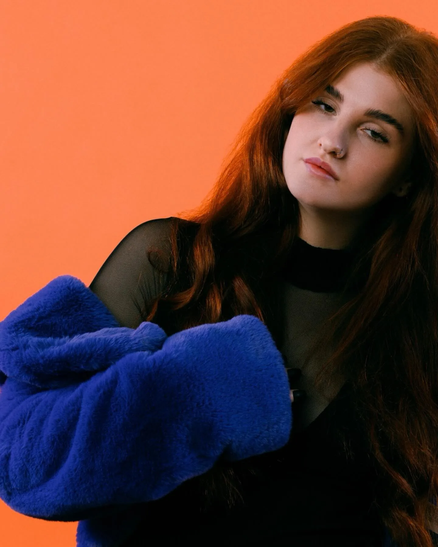 Daria against a flat orange studio backdrop in an electric blue faux-fur coat over sheer black mesh, chin lifted, long copper hair falling across the fur