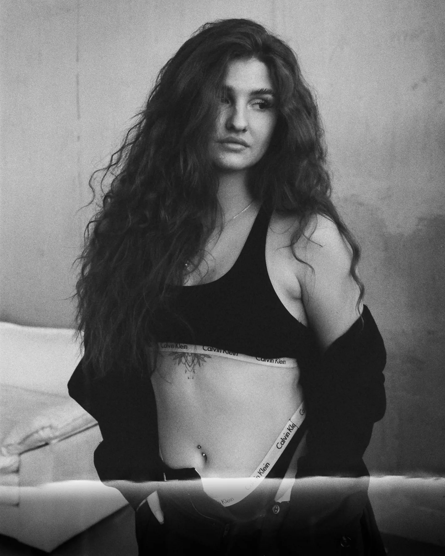Black-and-white portrait of Daria in a black blazer worn open over a bra and low jeans, dark curls falling forward, soft window light on a bare plaster wall