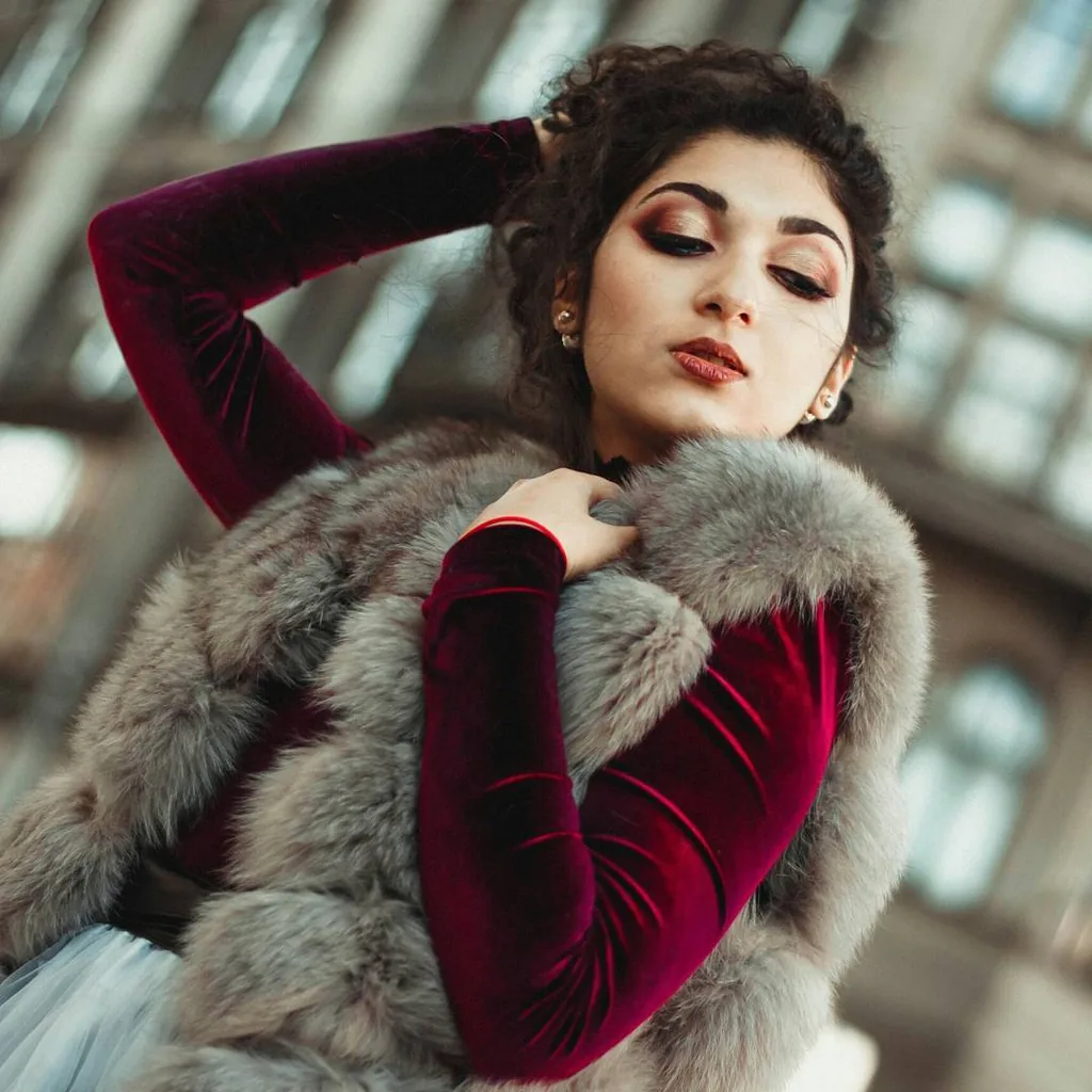 Model in a crimson velvet gown and grey fox fur stole, one arm raised into dark curls, eyes lowered, shot from below against a glass-and-iron arcade roof