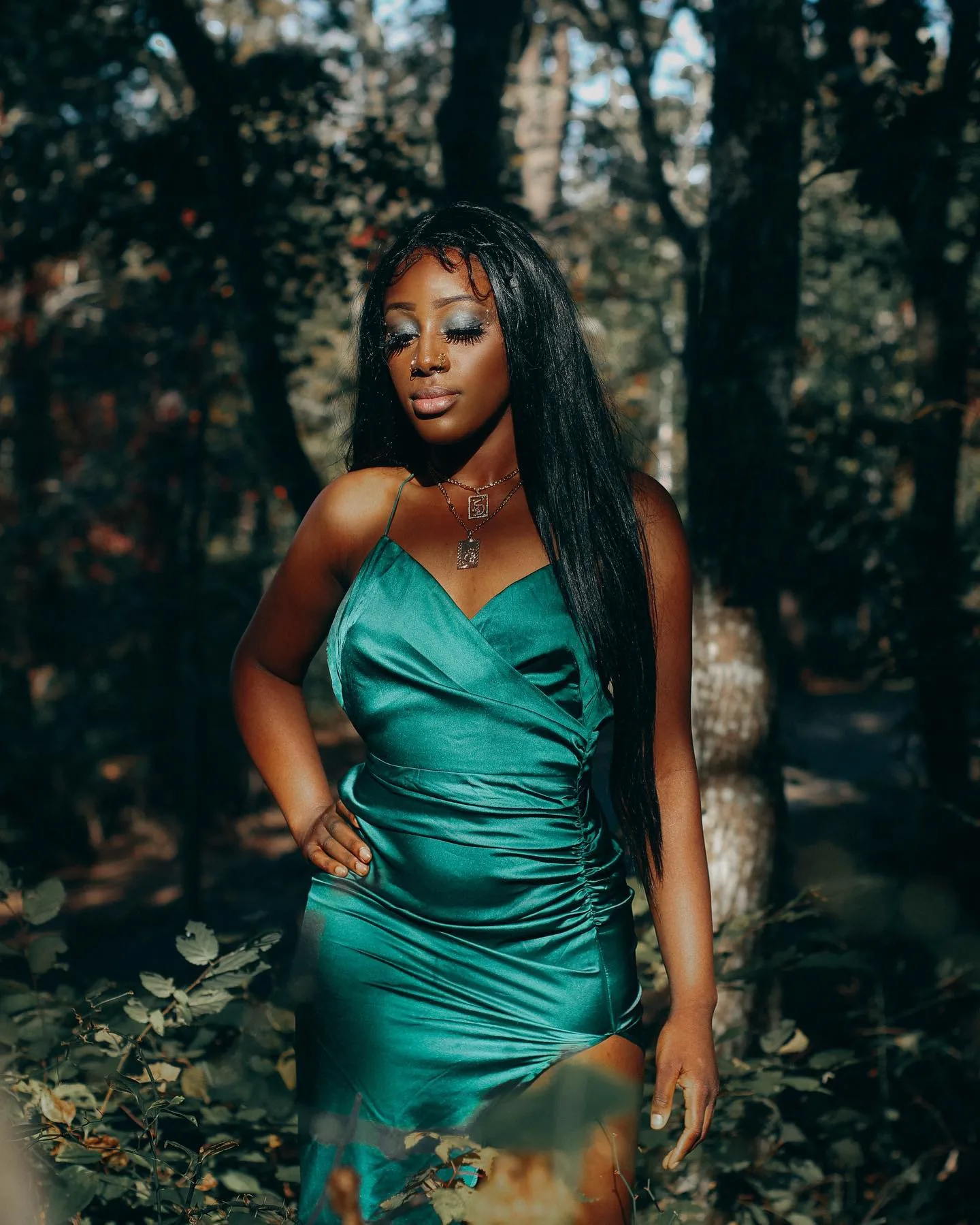 A woman in an emerald-green satin slip dress with a thigh split stands in autumn woodland, long dark hair falling past her waist, glitter eyeshadow catching the light, eyes lowered, hand on her hip in dappled late-afternoon sun.