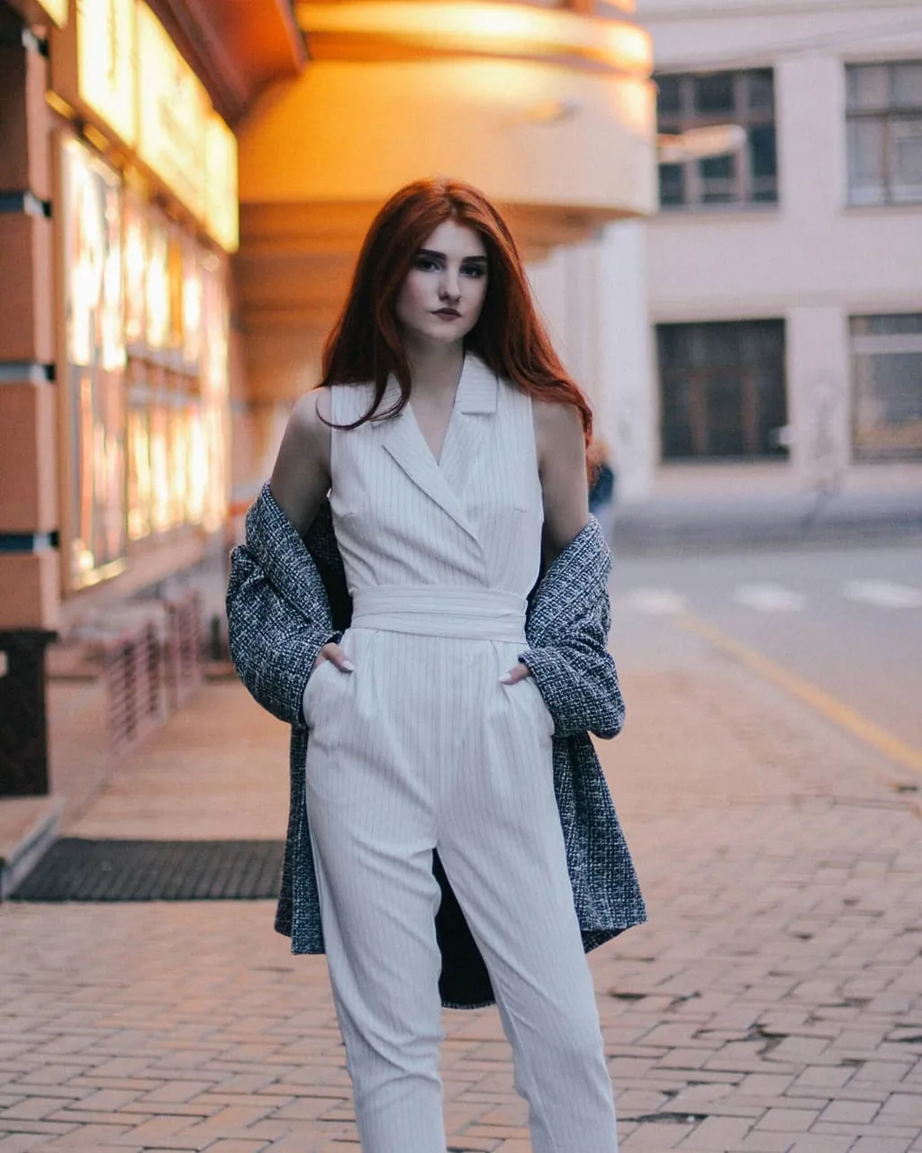 Full-length of Daria standing in an empty street at dusk in a white pinstriped sleeveless jumpsuit with a grey tweed coat slipped off the shoulders and hanging behind her, hands in pockets, warm lit shopfront glowing to one side.