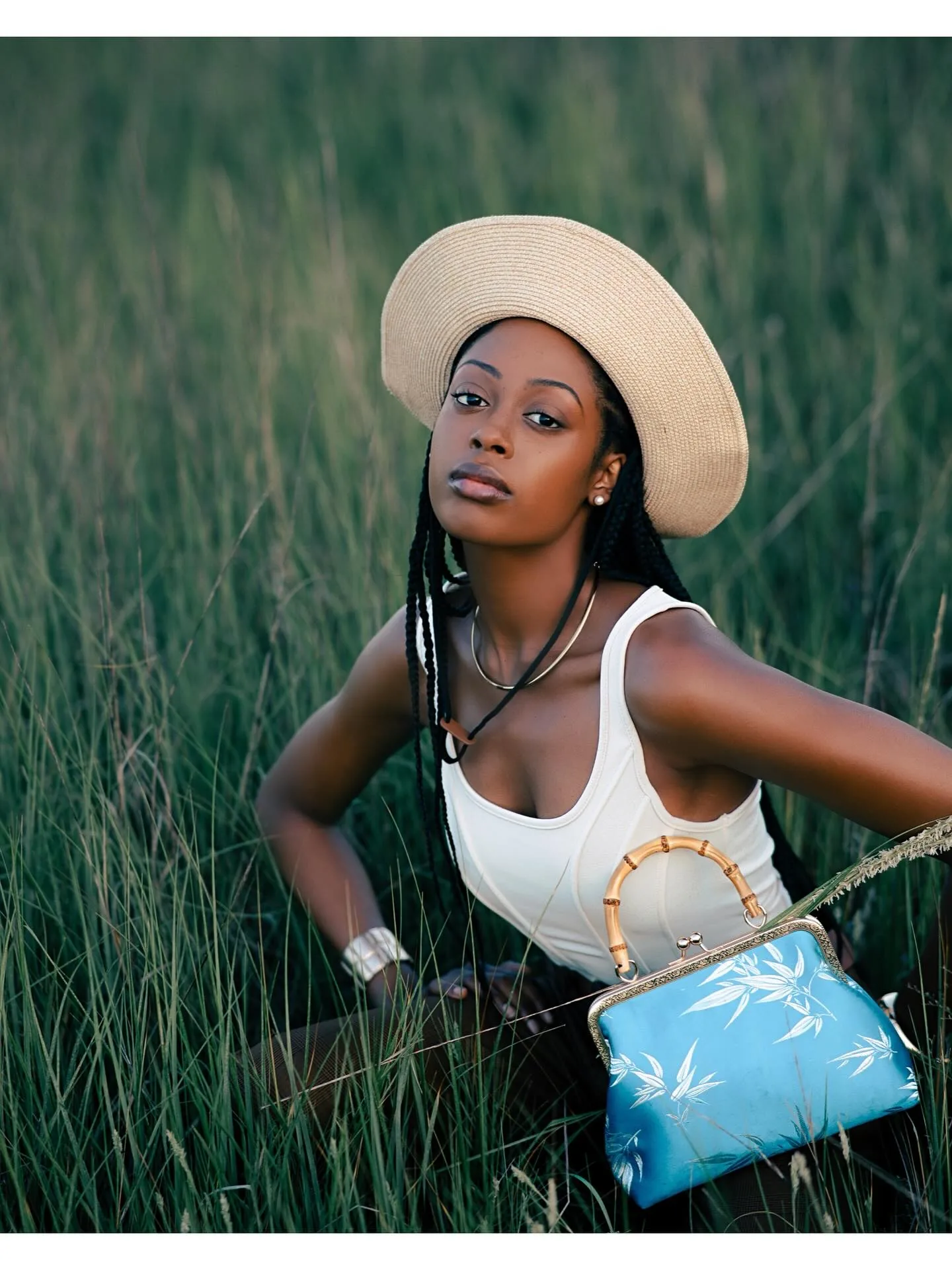 Golden-hour portrait of a model in a white tank top, brown belted trousers and a wide straw hat standing in tall marsh grass, hands on hips, holding a pale blue printed handbag.