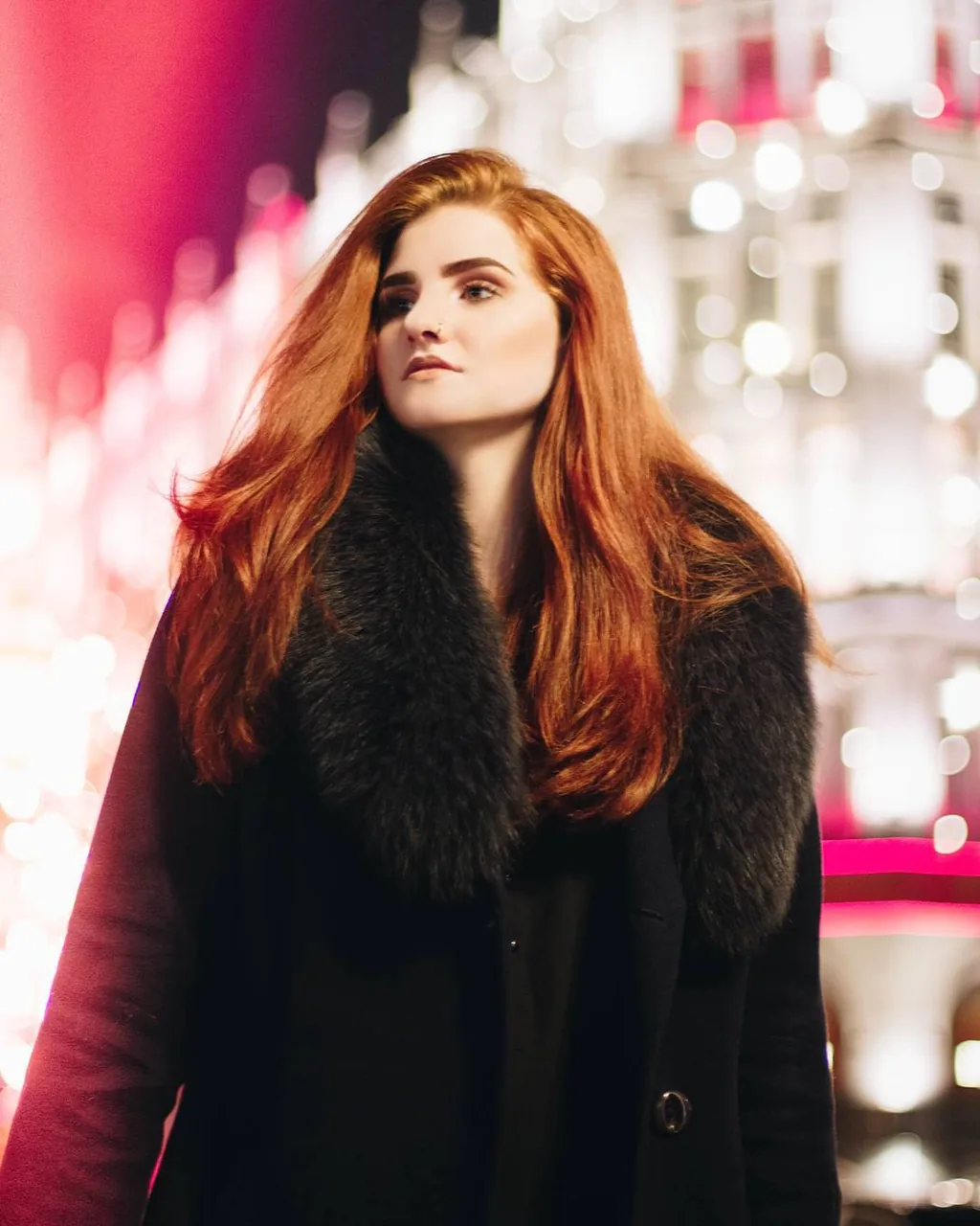 Daria on a Moscow street at night in a black coat with a deep black fur collar, copper hair falling loose, standing in front of a wall of pink and white out-of-focus city lights.