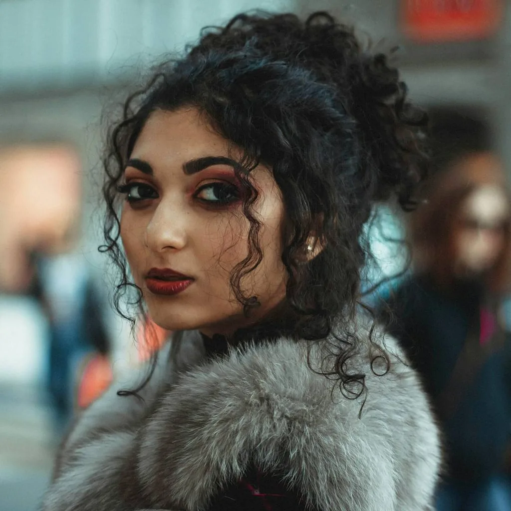 Close portrait of a model with dark curls piled high, deep red smoky eye and oxblood lip, glancing back over a silver fur collar, warm street bokeh behind