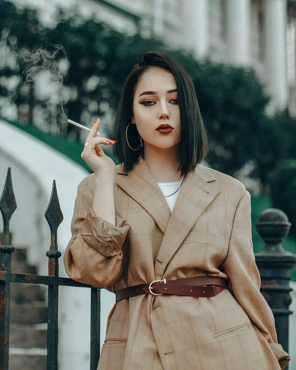 Portrait of a woman with a sharp dark bob, graphic orange eyeliner and deep red lip, wearing an oversized tan checked blazer cinched with a wide brown leather belt, holding a lit cigarette; wrought-iron railings and a pale classical facade behind her.