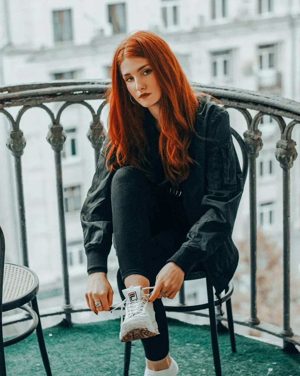 Daria on the same iron balcony, seated on a stool leaning forward to fasten a white chunky sneaker, black bomber and black jeans, red hair falling forward, pale facade behind.
