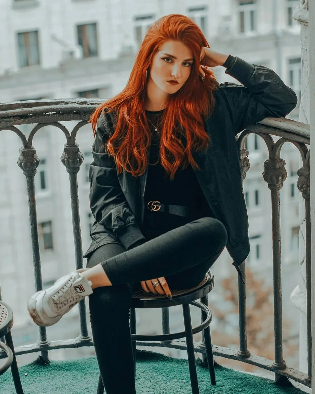 Daria seated on a bentwood stool on a wrought-iron European balcony, one leg drawn up, black bomber jacket over black tee and jeans with a gold GG buckle belt, white chunky sneakers, hand in her vivid orange-red hair, pale ornate facade behind.