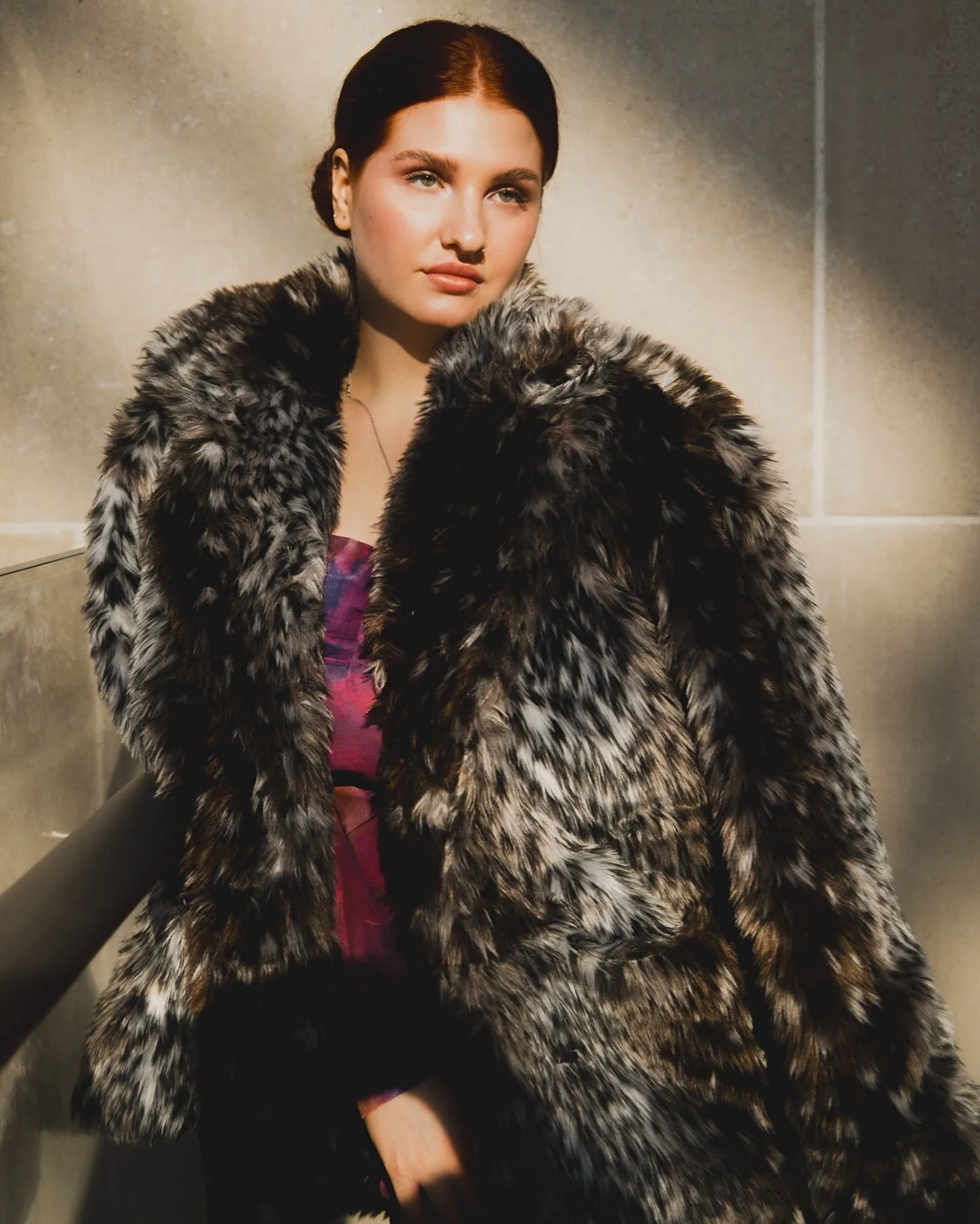 Daria seated on a marble staircase in a spotted black-and-white faux-fur coat over a magenta dress, an antique tapestry and stone column behind her, hard afternoon sun raking across the wall