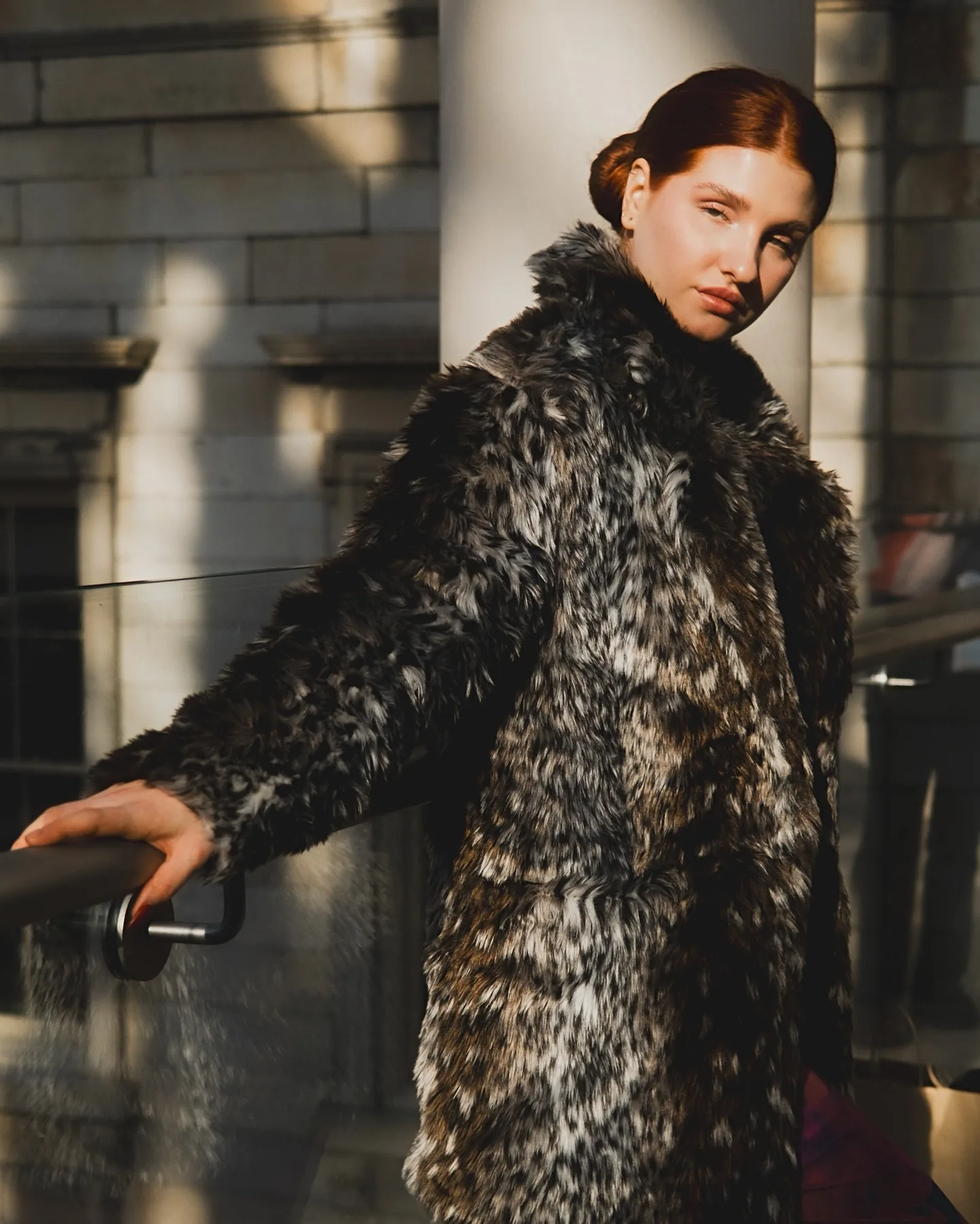 Daria in a spotted faux-fur coat leaning back with one hand along a stone stair rail, hair pulled tight, sunlight and window shadow falling in diagonal bands across pale limestone