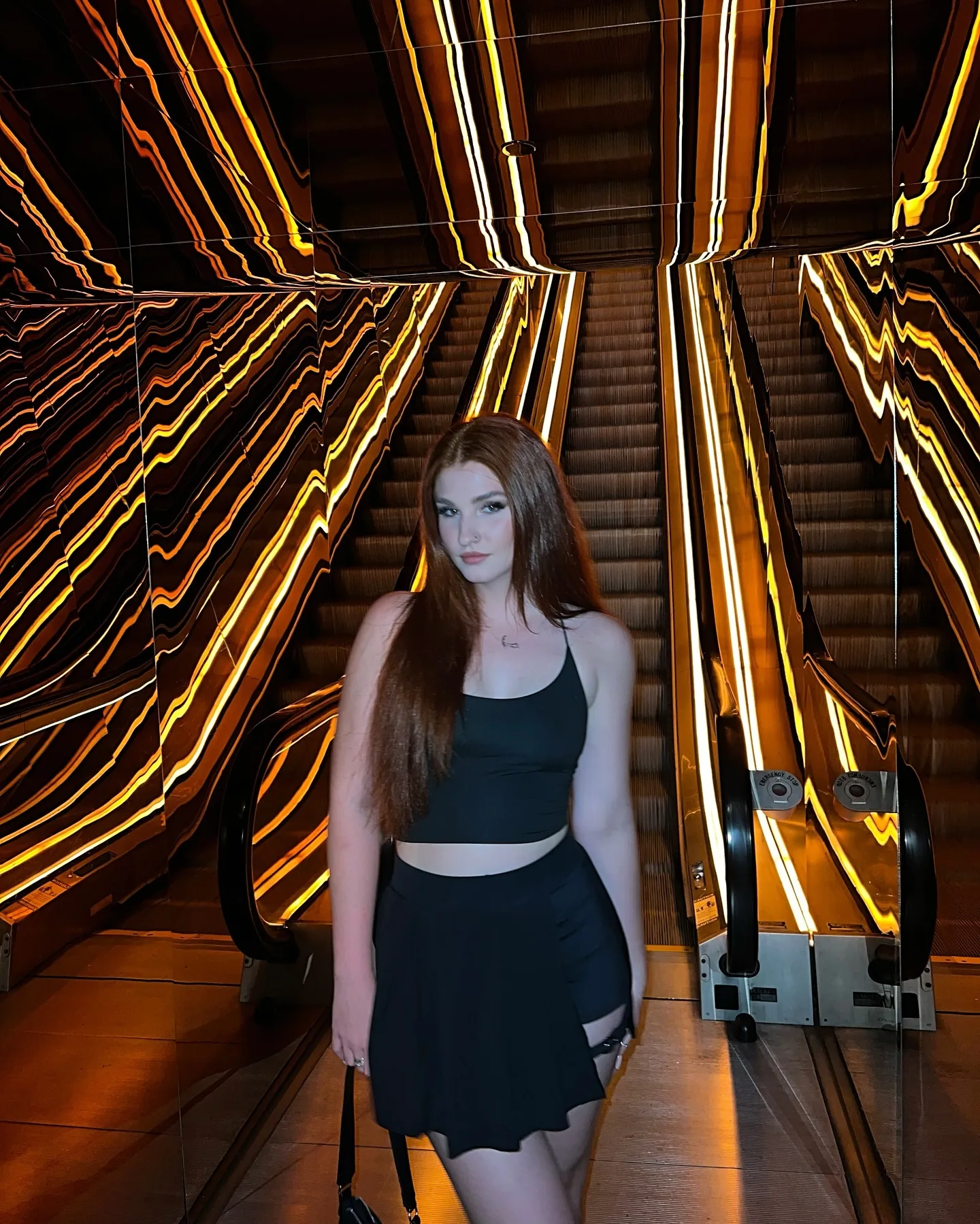 Daria standing at the foot of an escalator in a black crop top and short pleated skirt, long-exposure streaks of gold and amber light fanning out around her