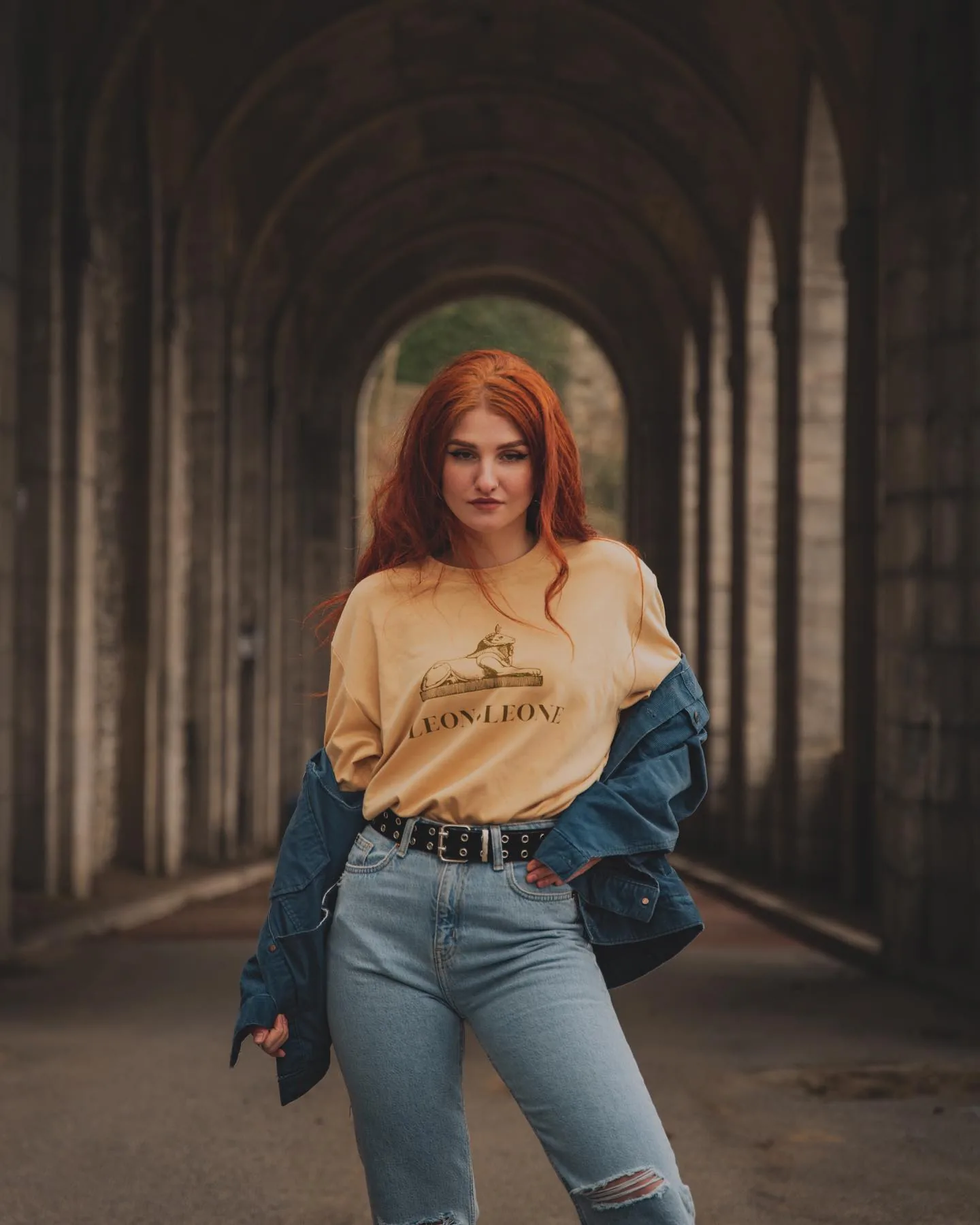 Woman with vivid red hair in an oversized butter-yellow graphic sweatshirt, denim jacket pushed off her shoulders, ripped light-wash jeans and a studded belt, standing in a long stone-vaulted arcade that recedes behind her.