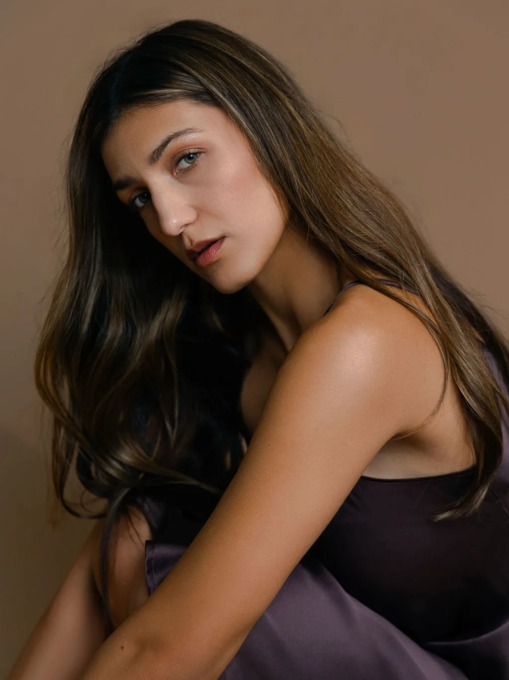 Warm-toned portrait of a long-haired woman in a plum silk slip dress seated against a tan backdrop, shoulder turned to camera, hair falling across her arm.