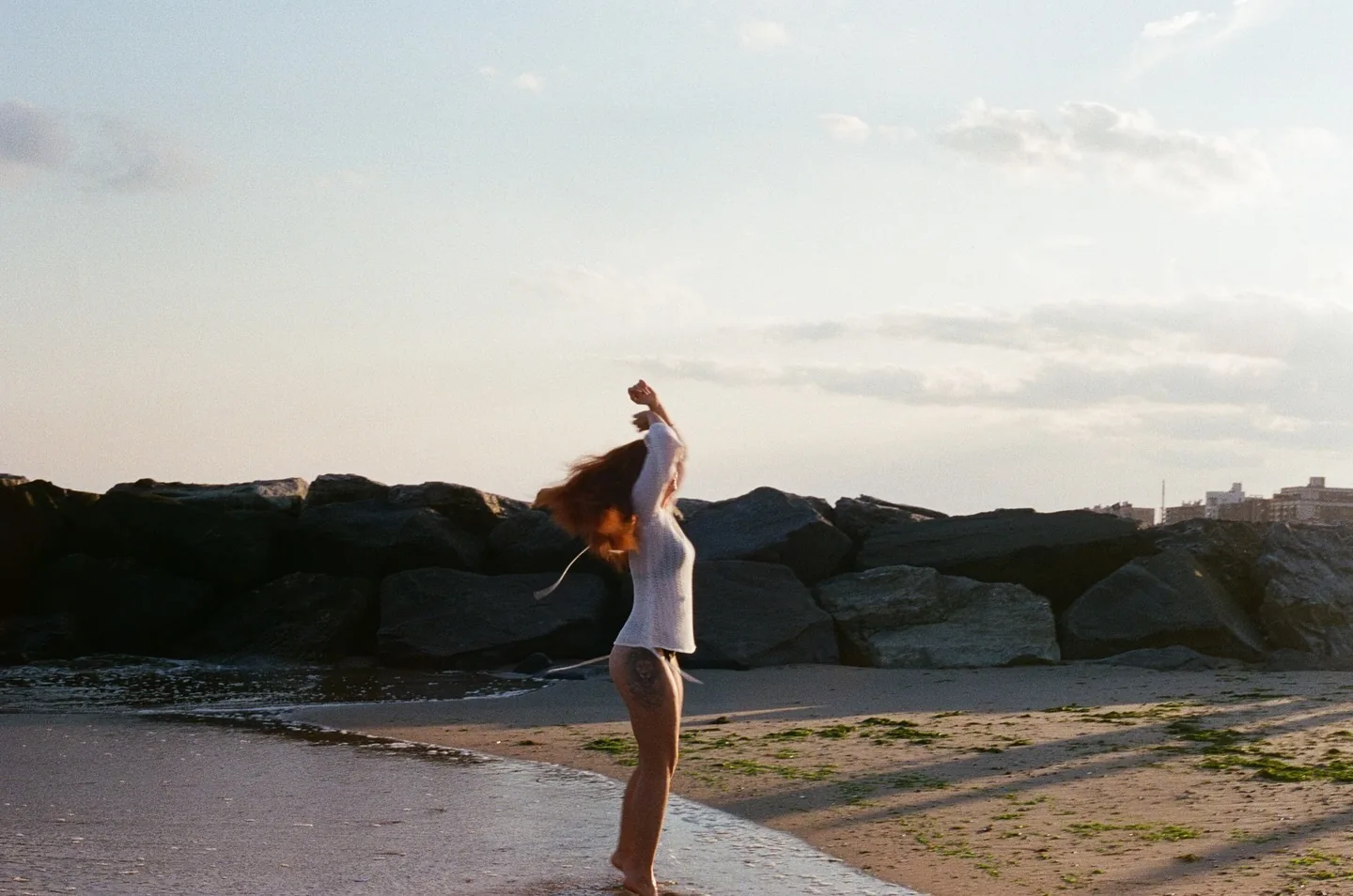 Backlit figure of Daria on the Coney Island sand at golden hour, arm thrown overhead and hair whipped across her face, a long dark stone jetty running behind her toward a pale sunset sky.