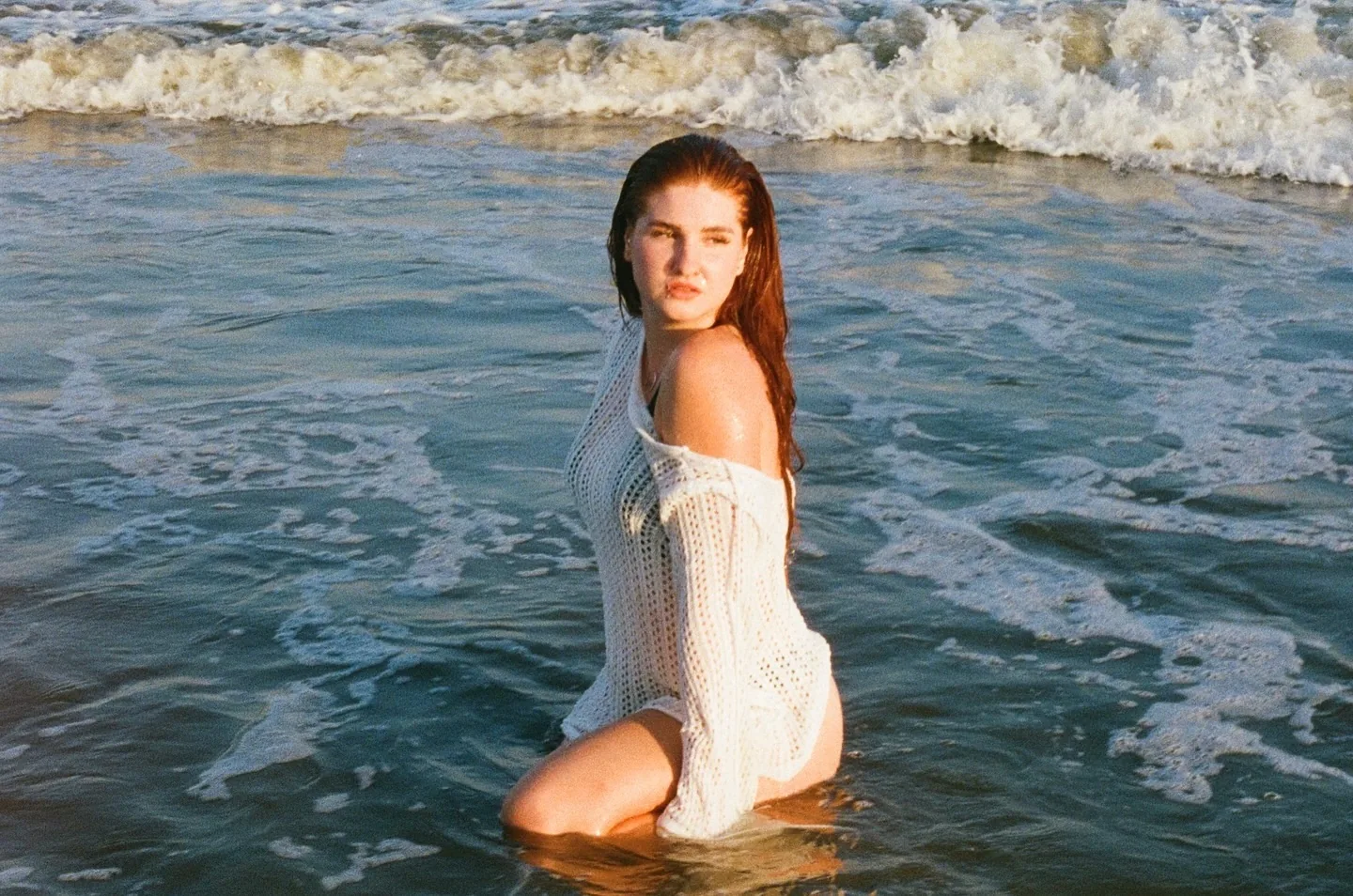 Daria kneeling waist-deep in the surf at Coney Island in a wet white crochet dress, hair slicked back, a large breaking wave folding into white foam directly behind her; shot on film with soft warm evening light on the water.