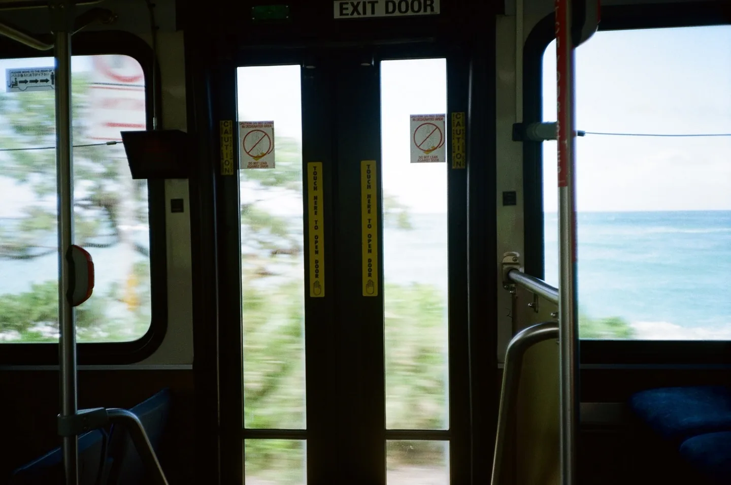 Interior of a moving Hawaii bus shot on film: dark rear doors and chrome poles in the foreground, roadside greenery streaked into motion blur and a band of turquoise ocean through the windows.
